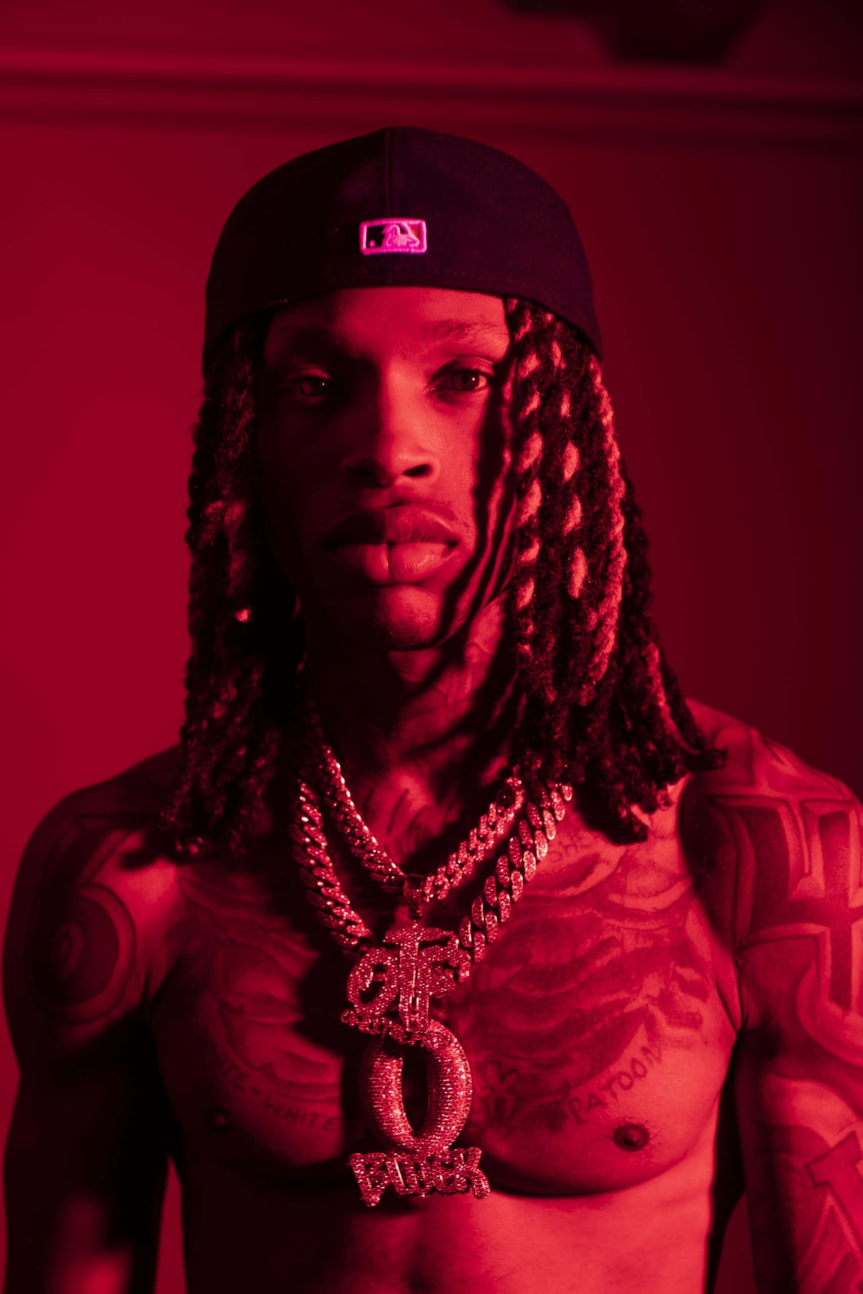 King Von 桌布 壁紙 — 一個赤裸上身、留著辮子頭和紋身的男人，在戲劇性的紅光下戴著棒球帽和沉重的鑲鑽鏈條，眼神銳利。