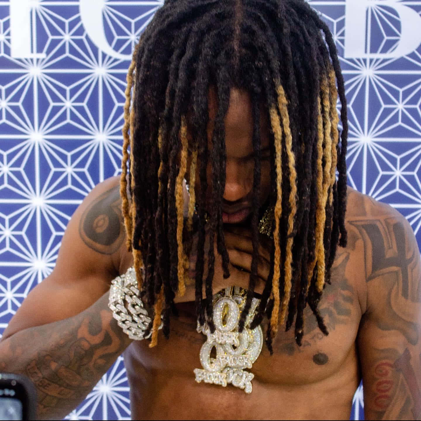 King Von 桌布 壁紙 — 一個赤裸上身的男人，留著黑色和金色的辮子頭，身上有紋身，戴著一條鑲滿鑽石的大鏈條，背景是藍色幾何圖案牆壁。