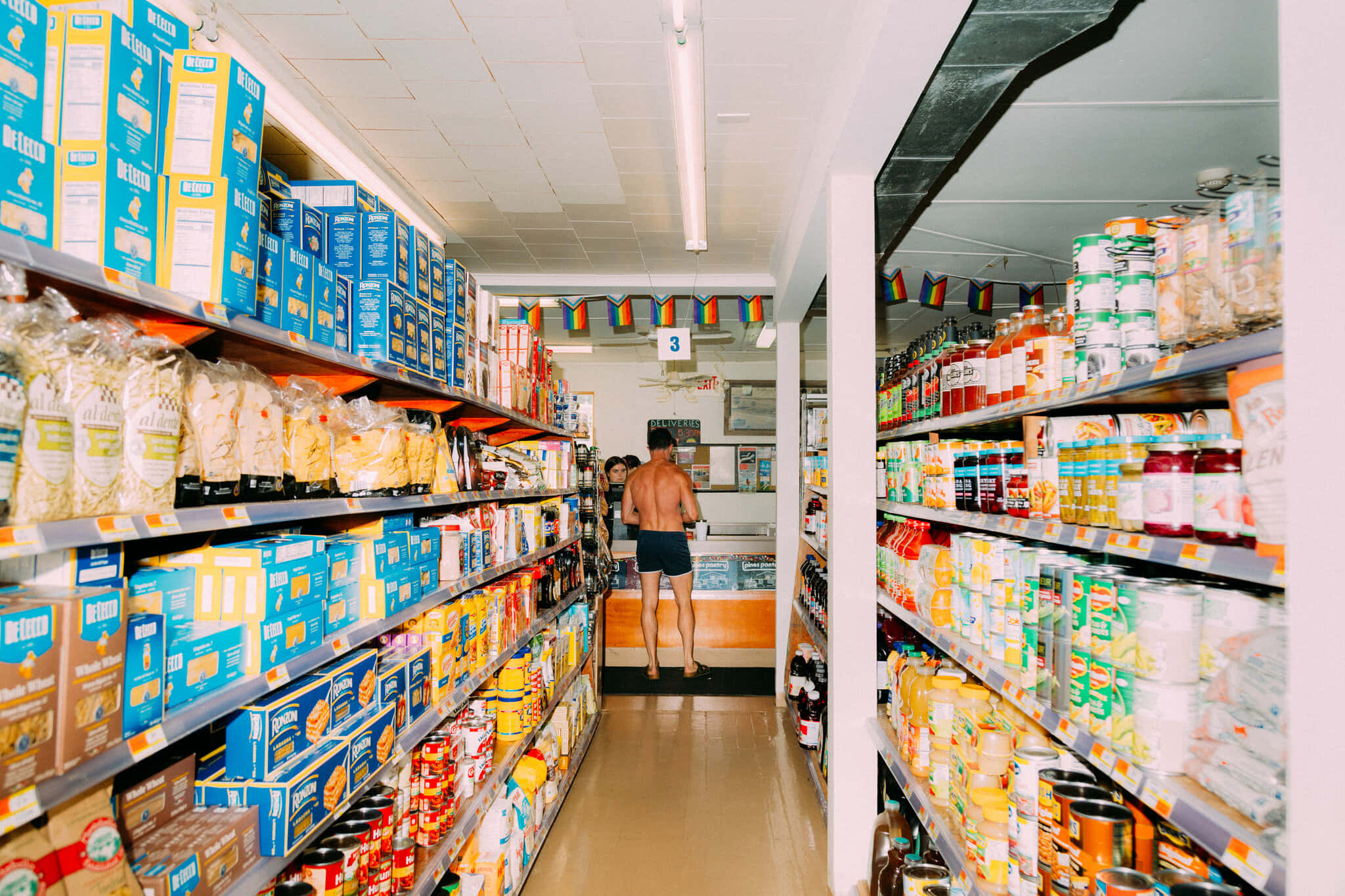 Grocery Store 桌布 壁紙 — 一個赤裸上身的男人穿著深色短褲，走在一條燈火通明的雜貨店通道上，兩旁貨架擺滿色彩繽紛的包裝商品。