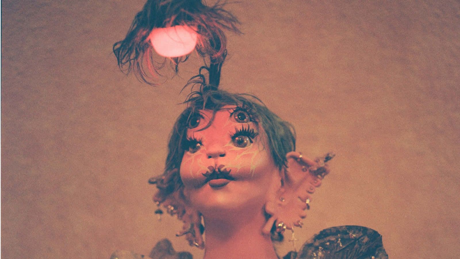Melanie Martinez 桌布 壁紙 — 一個超現實的肖像，描繪一個像玩偶的人物，有著誇張的眼睛和鬍子，頂部有一個發光的球體和深色羽毛，背景是柔和的棕色。