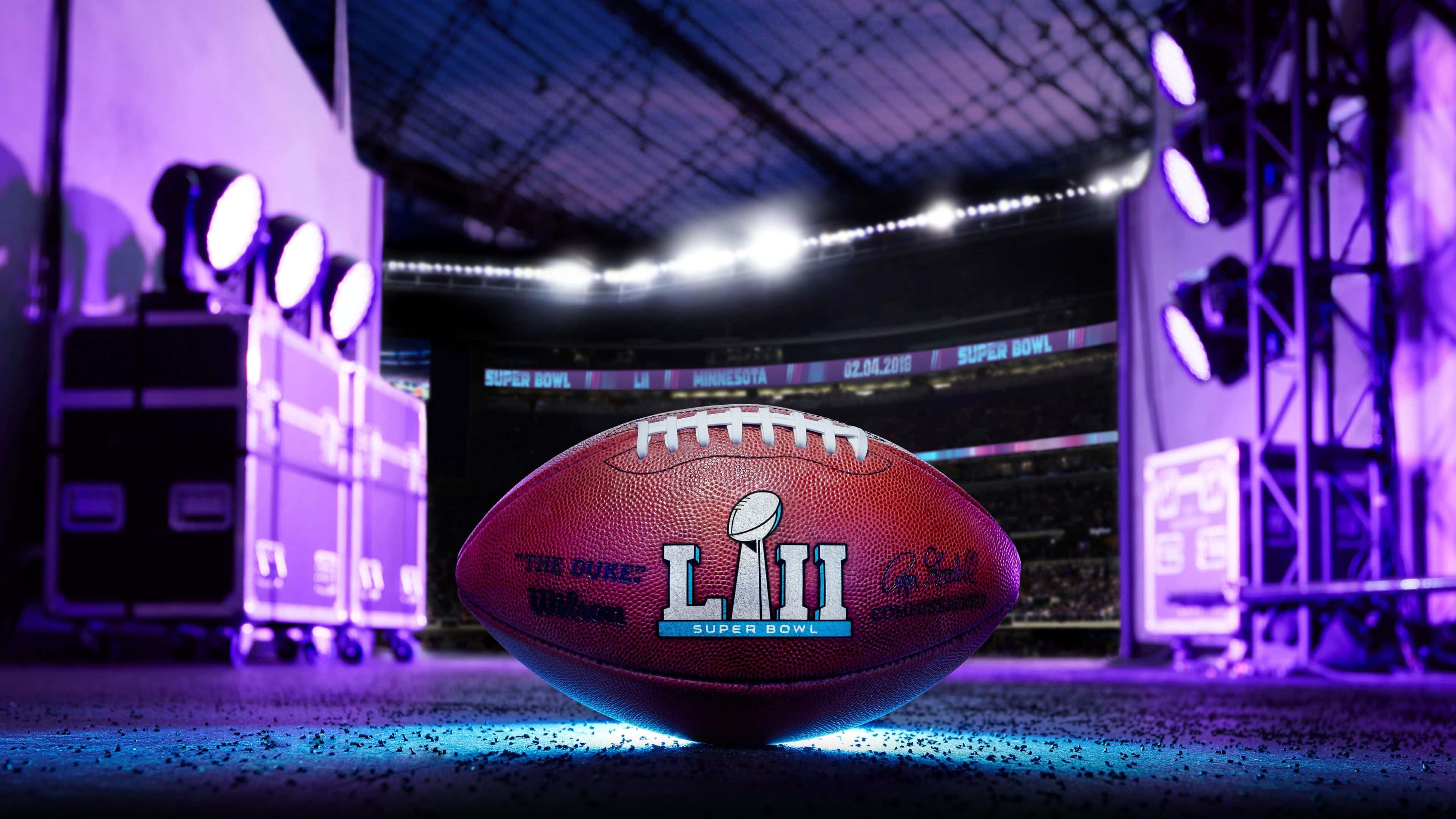 Super Bowl 桌布 壁紙 — 一個超級碗LII美式足球放在發光的藍色場地上，紫色體育場燈光下，背景是空無一人的看台。