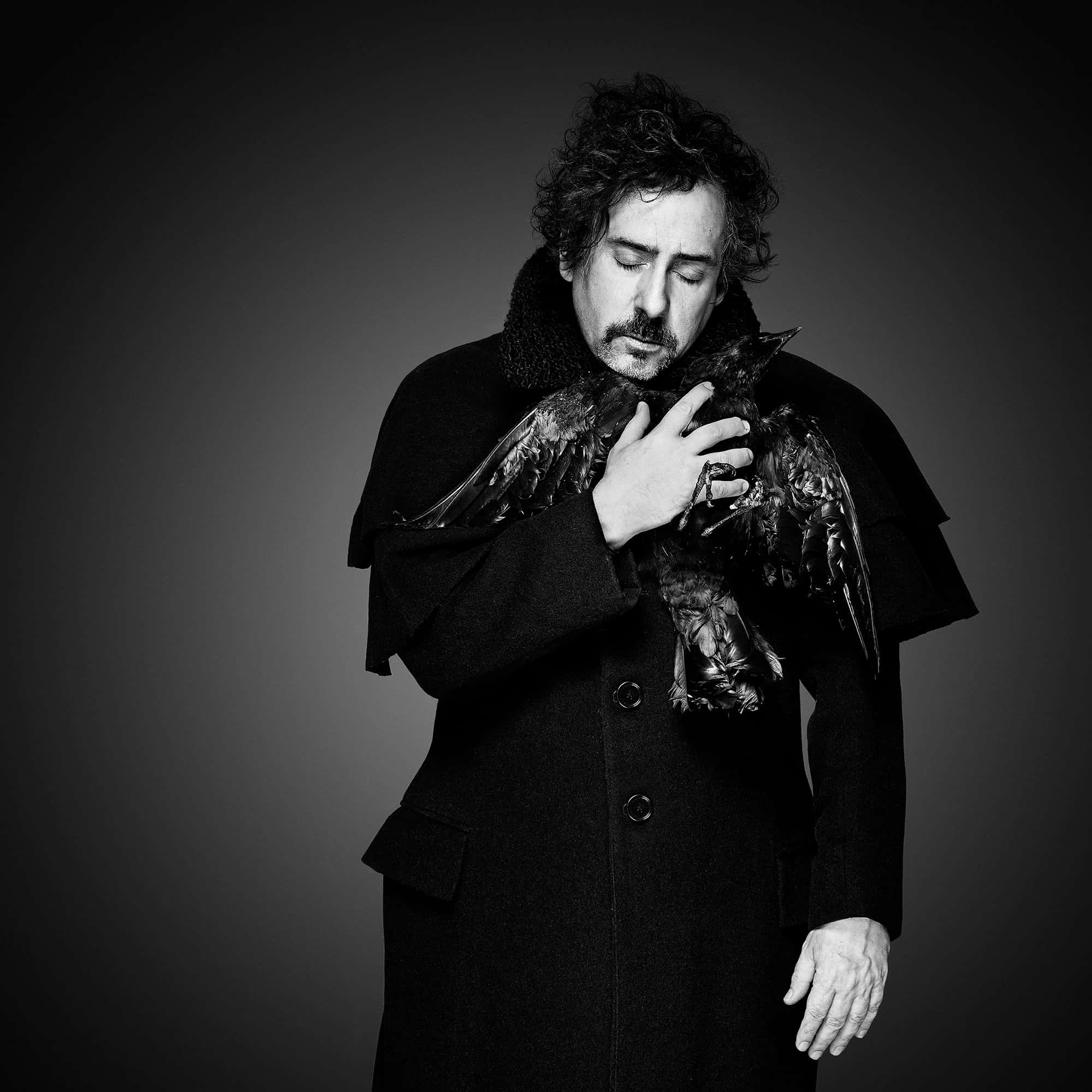 tim burton 桌布 壁紙 — 一個身穿深色外套的男人，在灰色漸變背景前輕輕懷抱一隻烏鴉，營造出陰鬱的黑白肖像。