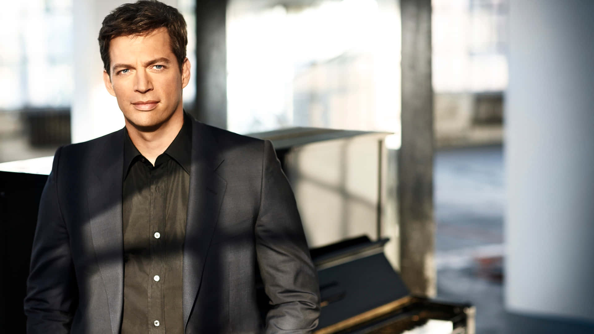 Harry Connick Jr 桌布 壁紙 — 一個身穿深色西裝的男人自信地站在現代、陽光充足的房間裡，旁邊有一部三角鋼琴，房間有大窗戶。