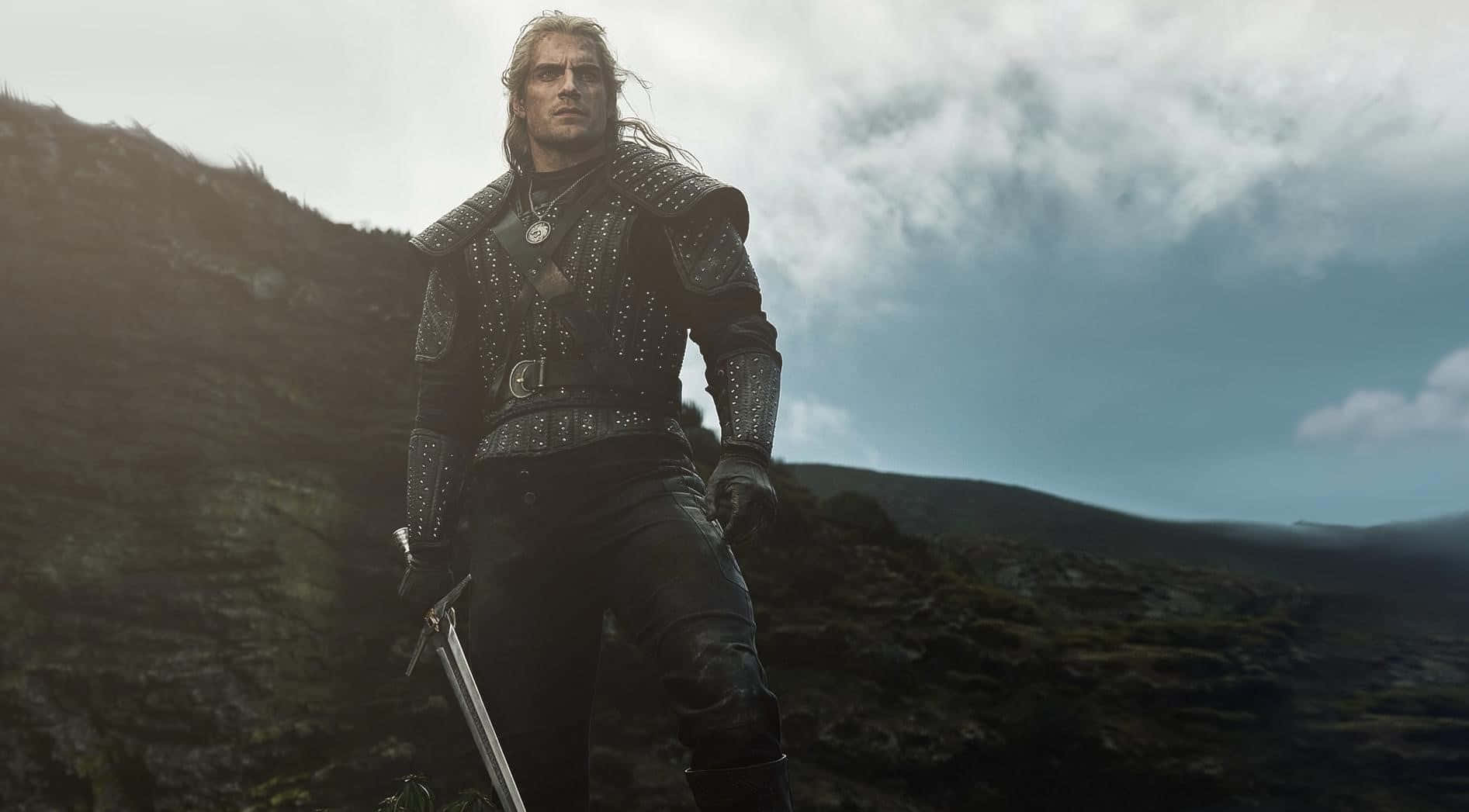 The Witcher Netflix 桌布 壁紙 — 一個身穿深色鉚釘盔甲的粗獷戰士，站在霧氣繚繞的山脊上，在陰雲密佈的天空下手持一把劍。