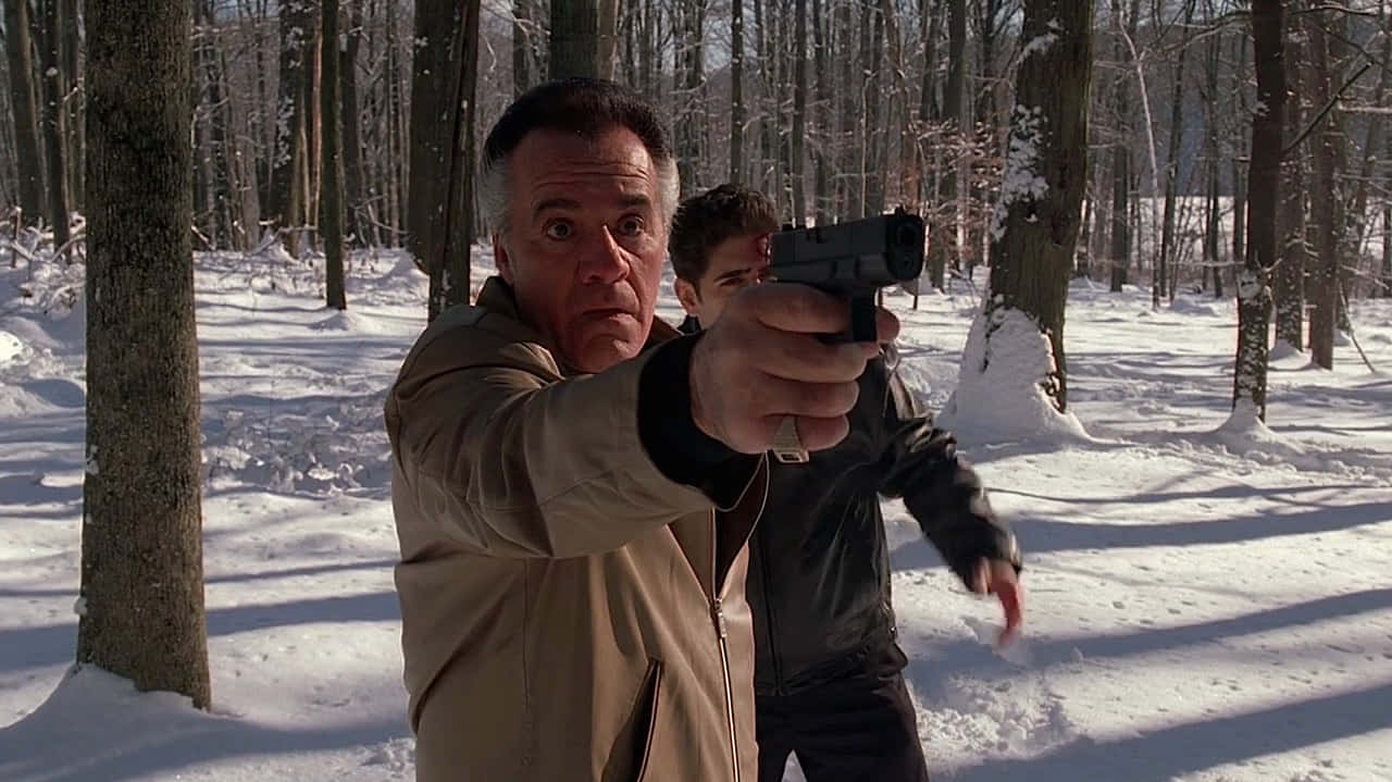 Tony Sirico 桌布 壁紙 — 一個身穿淺棕色外套的男人在雪地森林中向前瞄準手槍，身後有另一個男人。