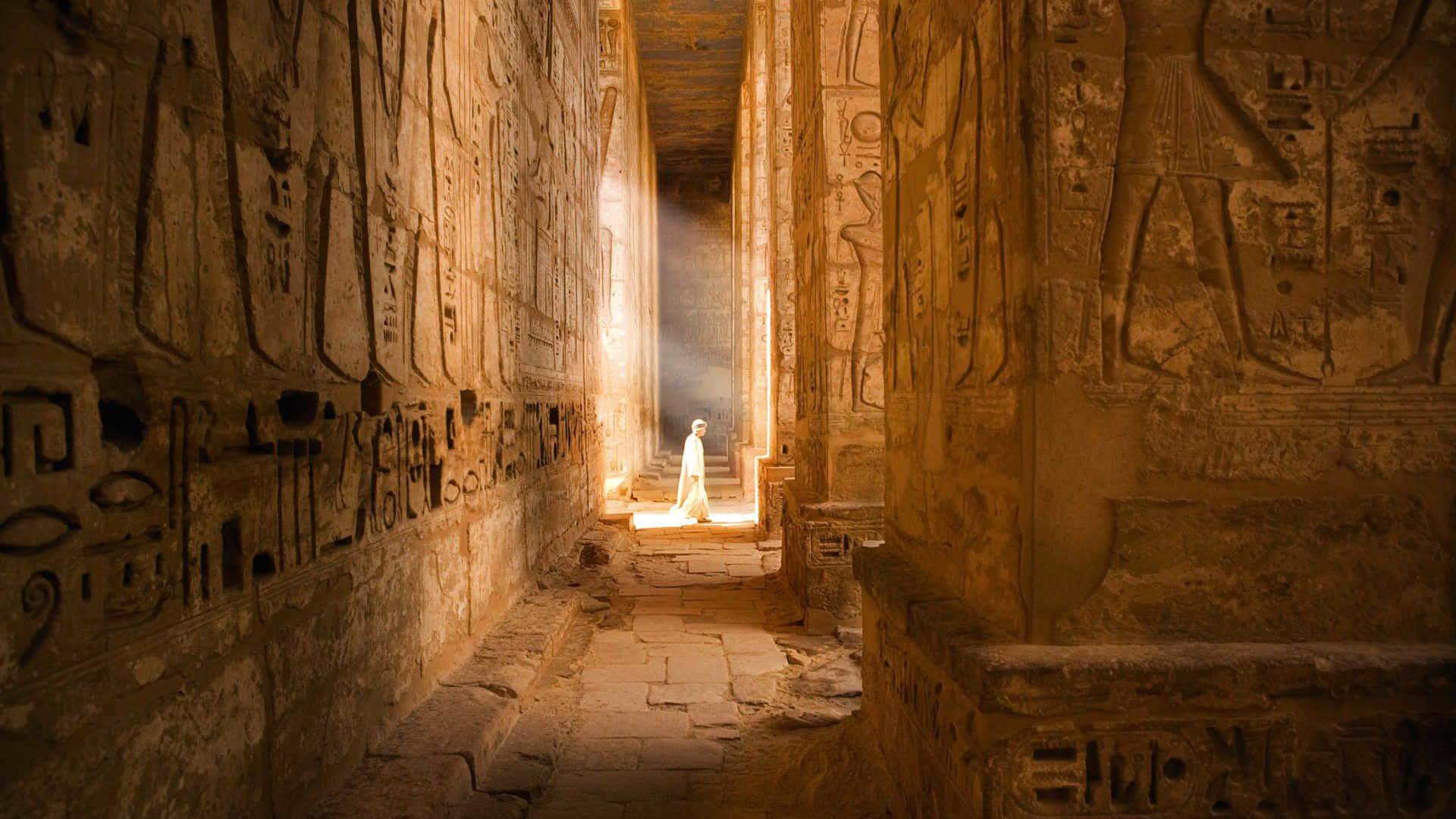 Egypt 桌布 壁紙 — 一個身穿白衣的孤獨身影，站在刻有象形文字和雕像的古埃及神廟走廊盡頭，沐浴在陽光下。