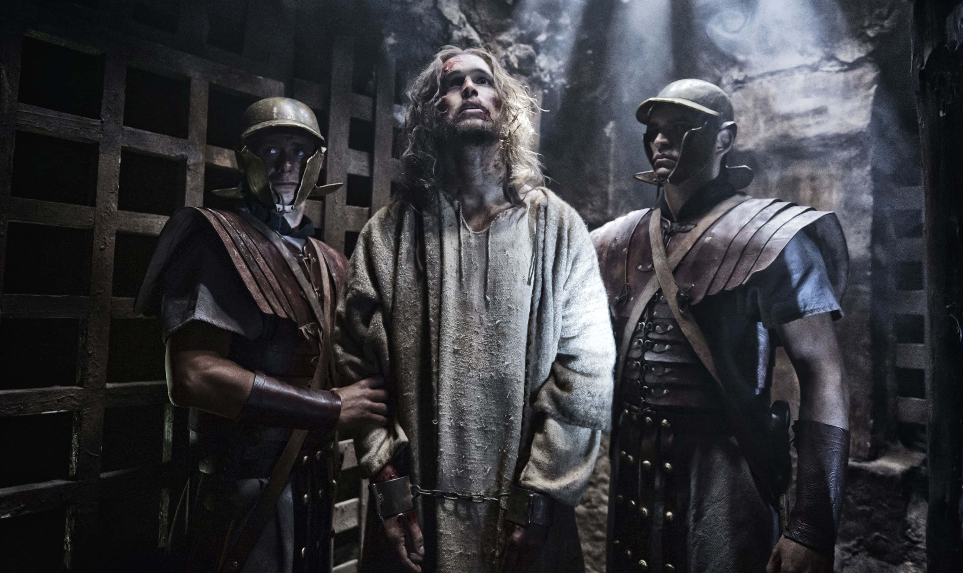 Son Of God 桌布 壁紙 — 一個身穿破爛長袍的男人站在兩名羅馬士兵之間，身處一個昏暗的石頭監獄，有鐵欄窗戶和閃爍的火把光。