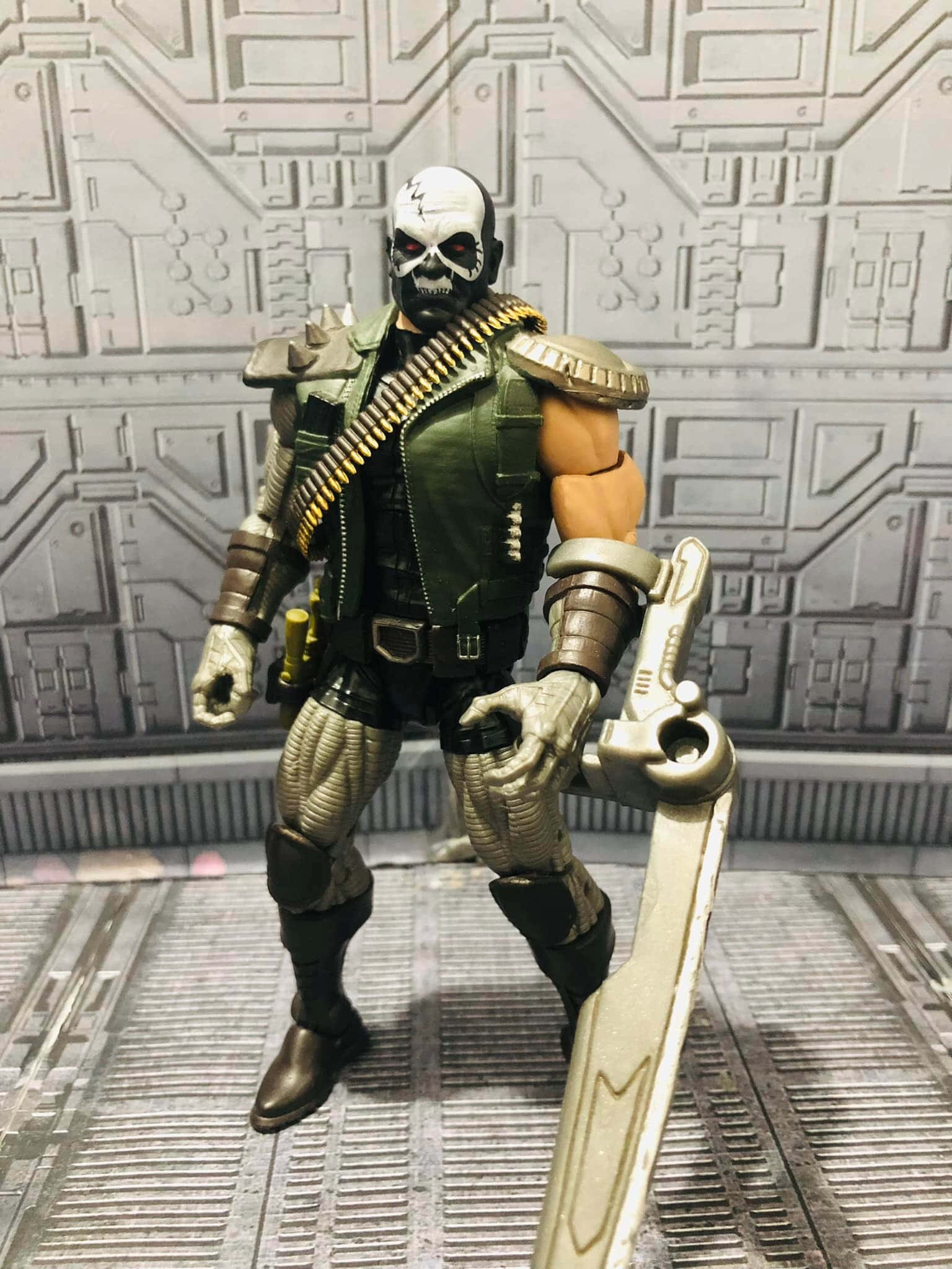Skullbuster 桌布 壁紙 — 一個身穿綠色盔甲、面目猙獰的骷髏頭動作人偶，站在金屬格柵地板上，背景是電路圖案牆壁，手持劍蓄勢待發。