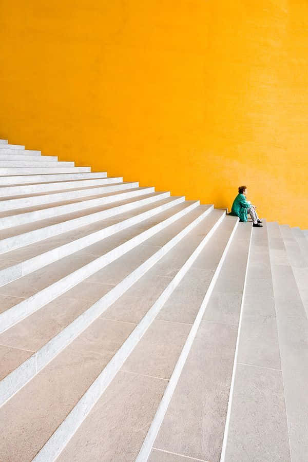 Minimalist Photography 桌布 壁紙 — 一個身穿綠色衣服的人獨自坐在寬闊樓梯的頂端，背景是鮮豔的橙色牆壁。