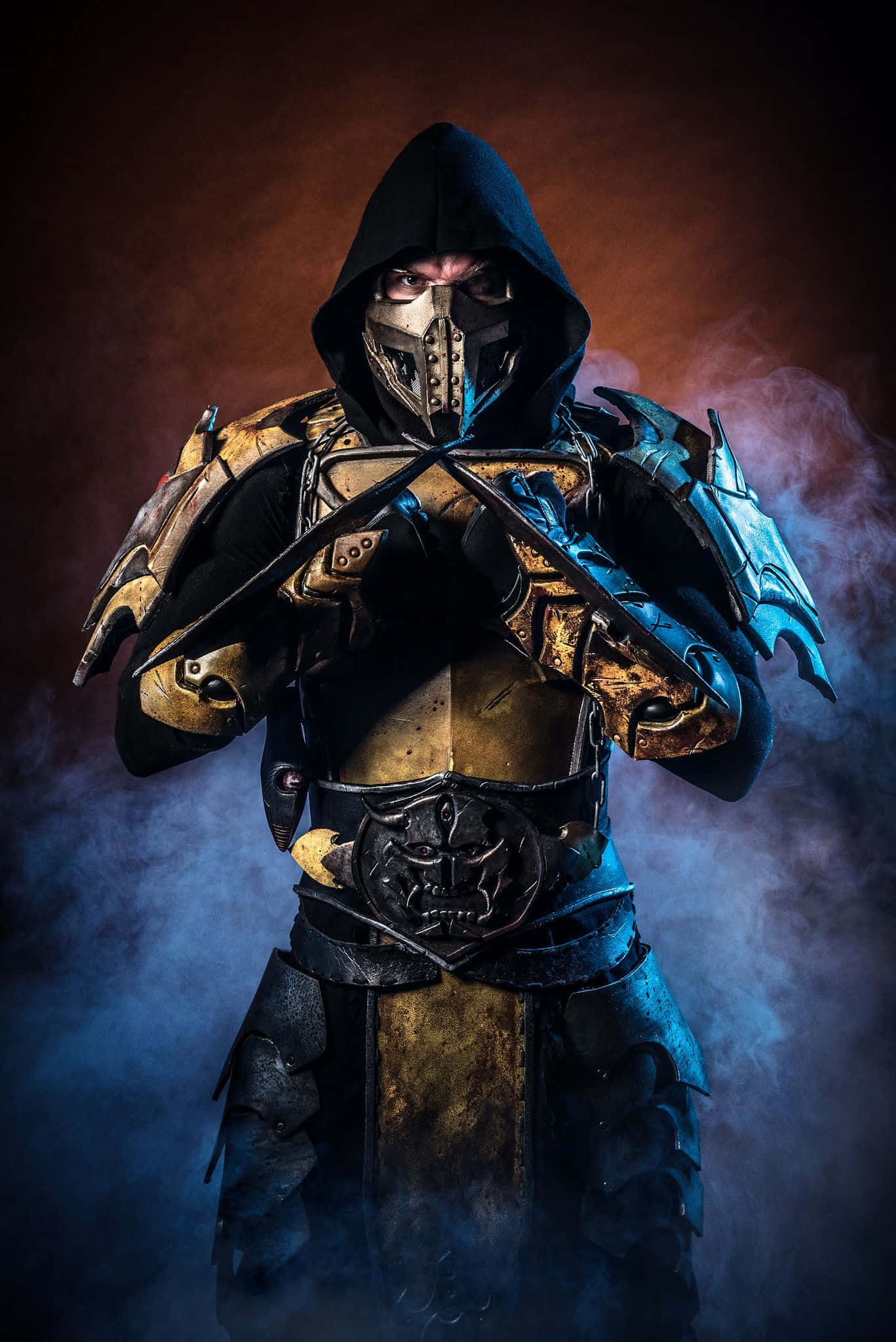 Mortal Kombat Cosplays 桌布 壁紙 — 一個身穿華麗金黑盔甲的威脅性兜帽戰士，在煙霧瀰漫的黑暗中站立，雙眼發光，手持交叉刀刃。
