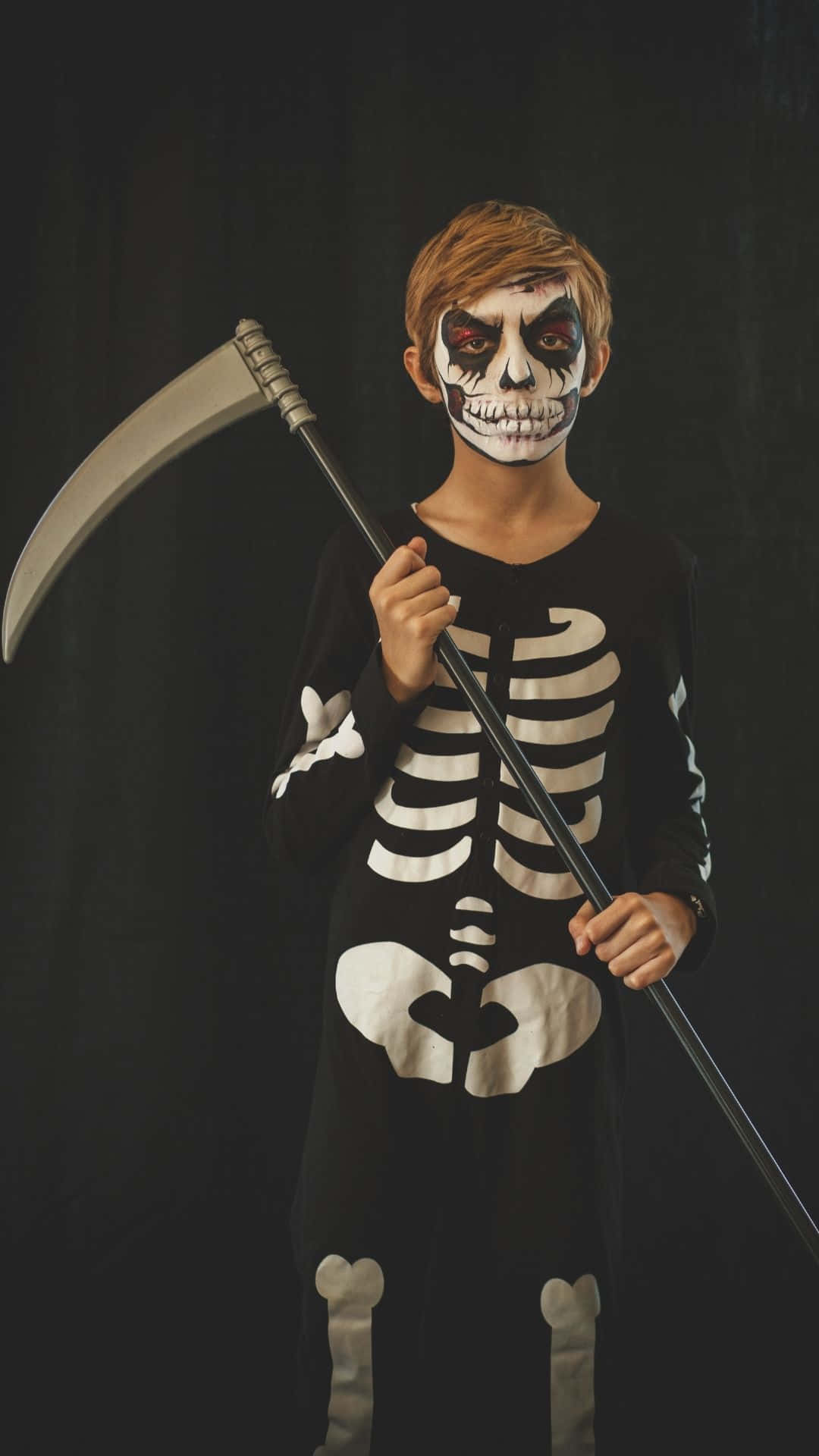 Skeleton Costumes 桌布 壁紙 — 一個身穿骷髏服裝、化著骷髏妝的小男孩，在黑暗背景下手持鐮刀。