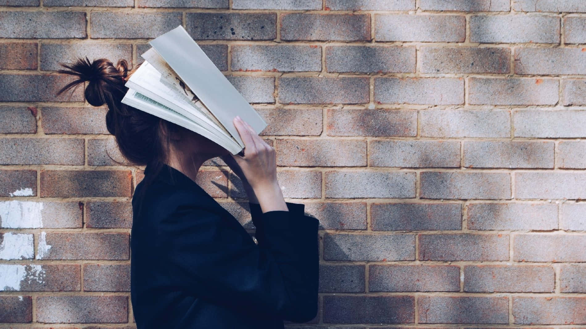 溫習 桌布 壁紙 — 一個身穿黑色外套的人用一本打開的書遮住臉，靠在一面風化的磚牆上。