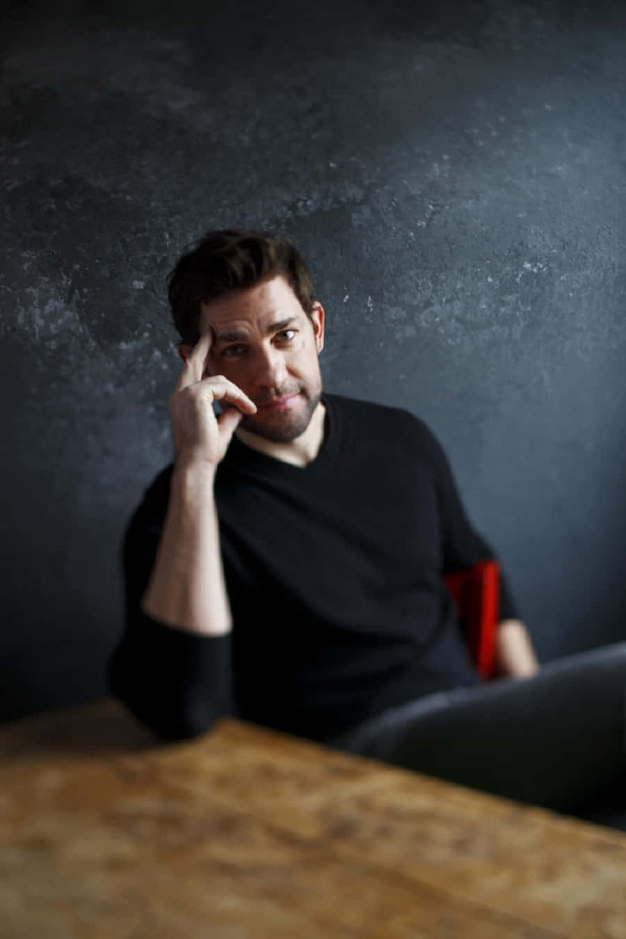 john krasinski 桌布 壁紙 — 一個身穿黑色恤衫嘅男人坐喺木枱前，背景係深色有紋理嘅牆，手放喺太陽穴，若有所思。