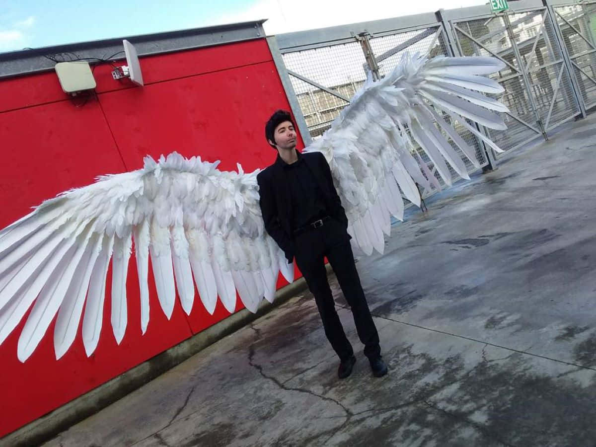 Lucifer Wings 桌布 壁紙 — 一個身穿黑色西裝的男人自信地站著，背後有巨大的白色天使翅膀，背景是紅牆和金屬圍欄。