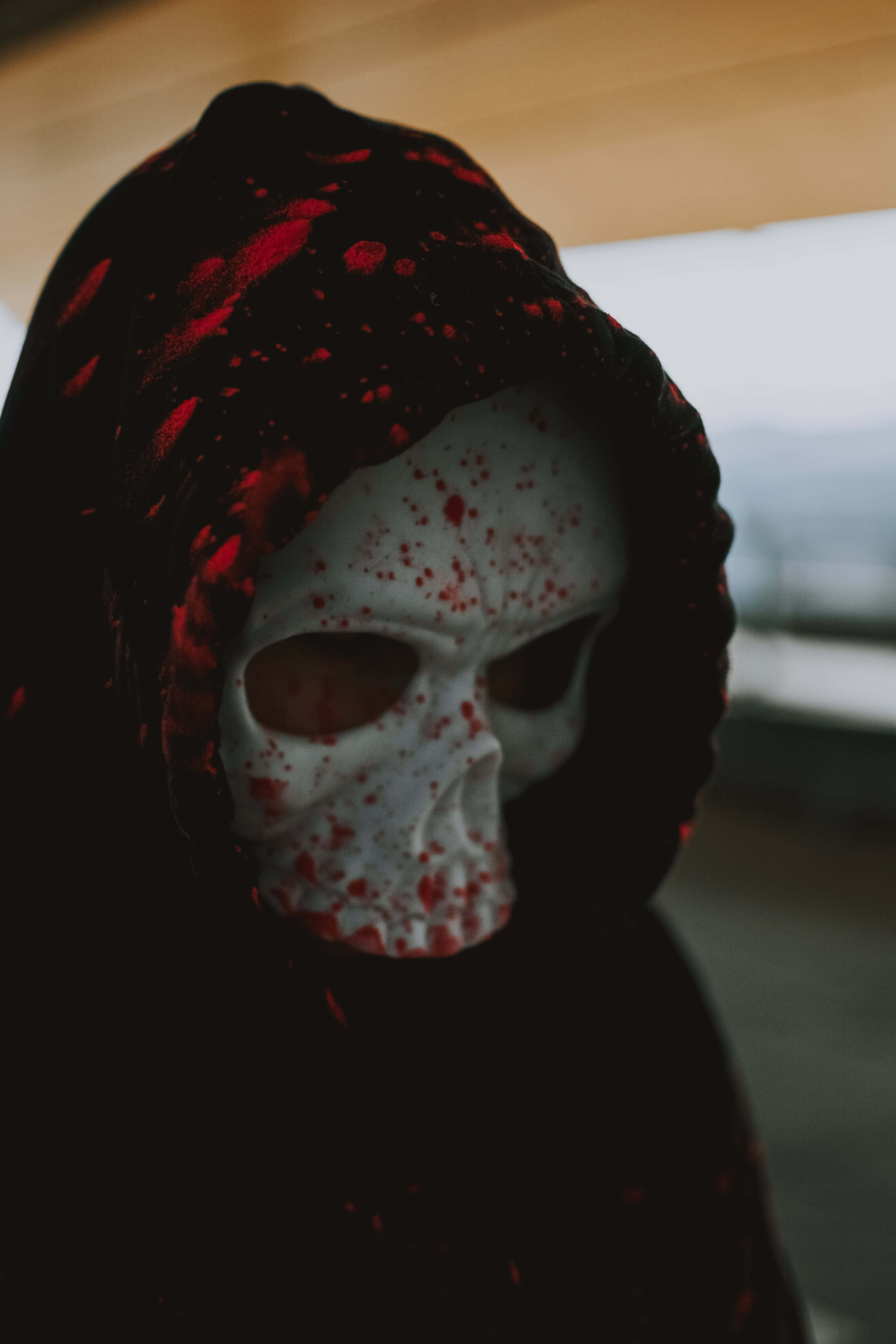 Horror Masks 桌布 壁紙 — 一個身穿黑色連帽斗篷、帶有紅色濺污的人物，戴著沾有血跡的白色骷髏面具，背景是模糊的城市景觀。