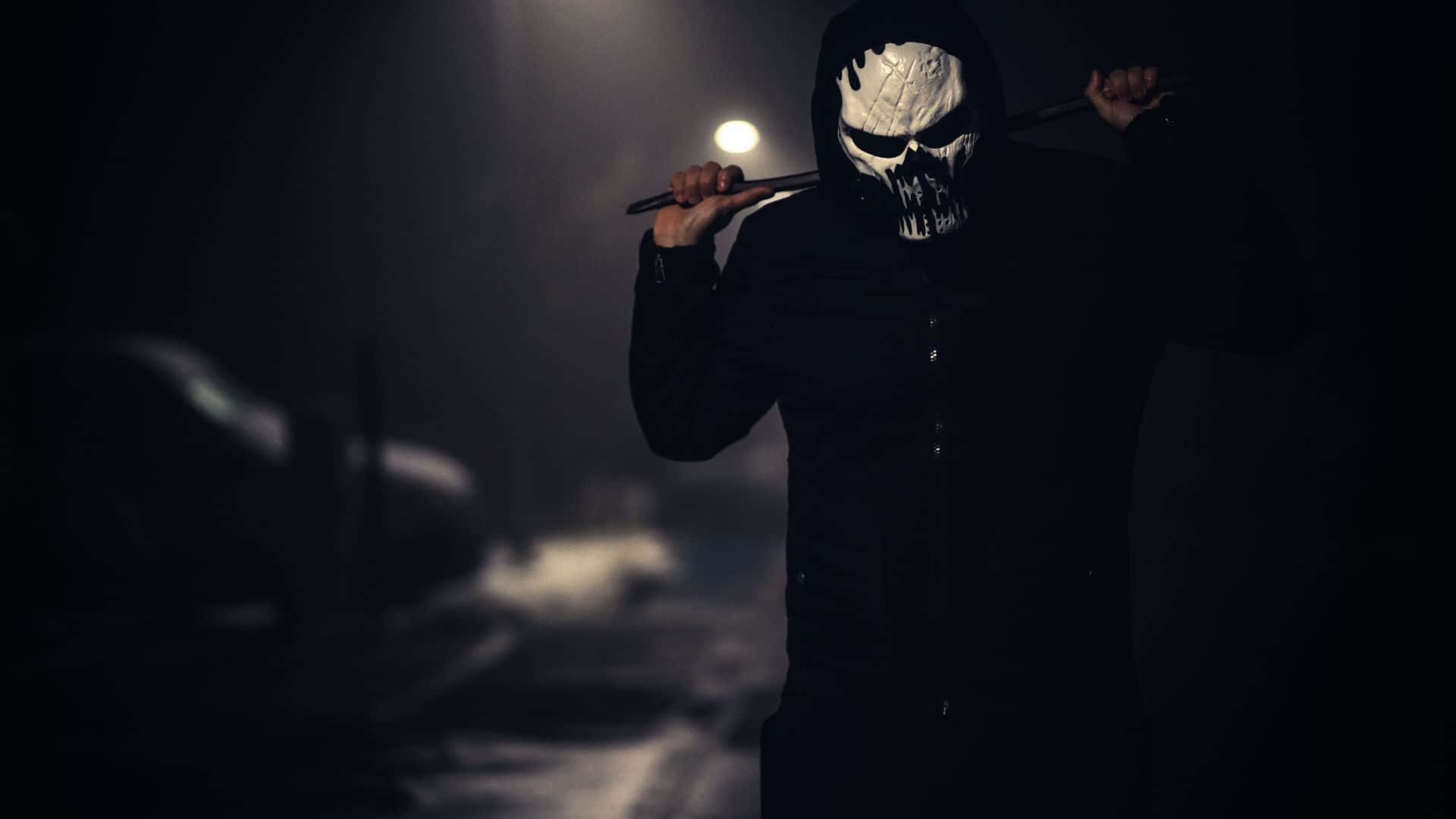 Horror Costumes 桌布 壁紙 — 一個身穿黑色連帽衫、戴著白色骷髏面具的人物，在煙霧瀰漫的黑暗小巷中，於單一聚光燈下手持警棍。