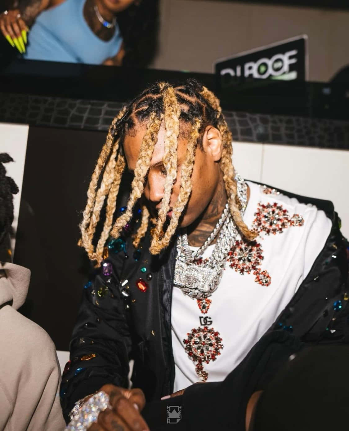 lil durk 桌布 壁紙 — 一個辮子頭髮、戴著閃亮珠寶的男人在燈光昏暗的俱樂部環境中向前傾身。