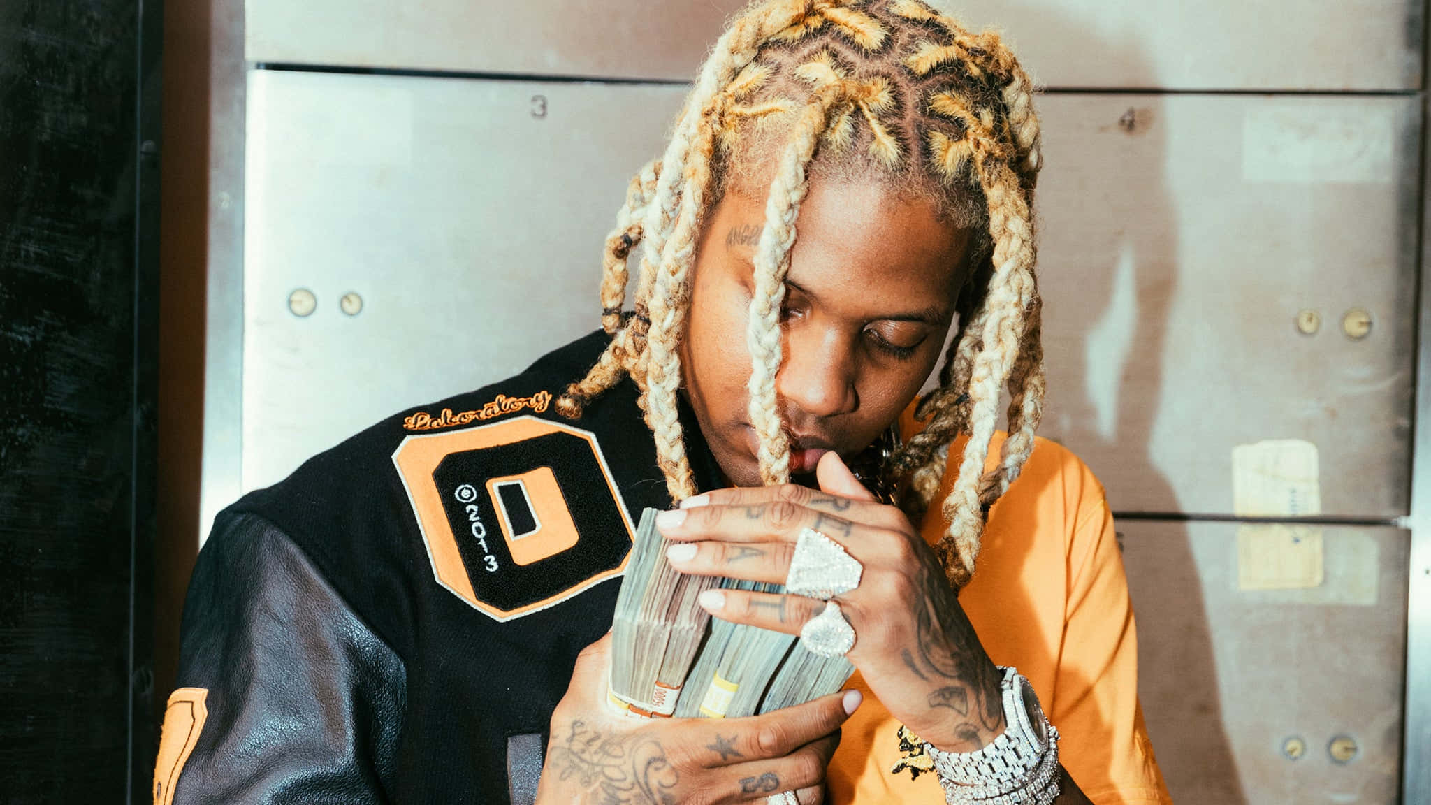 lil durk 桌布 壁紙 — 一個辮子頭髮的人穿著黑色和橙色外套，在金屬更衣室裡拿著一疊現金，低頭看著。