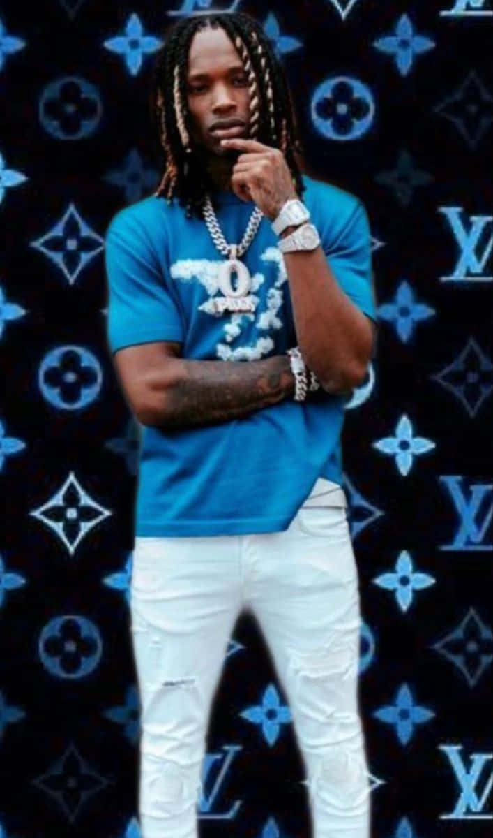 King Von 桌布 壁紙 — 一個辮子頭髮的男人，穿著藍色T恤和白色褲子，在黑色Louis Vuitton圖案背景前若有所思地擺姿勢。