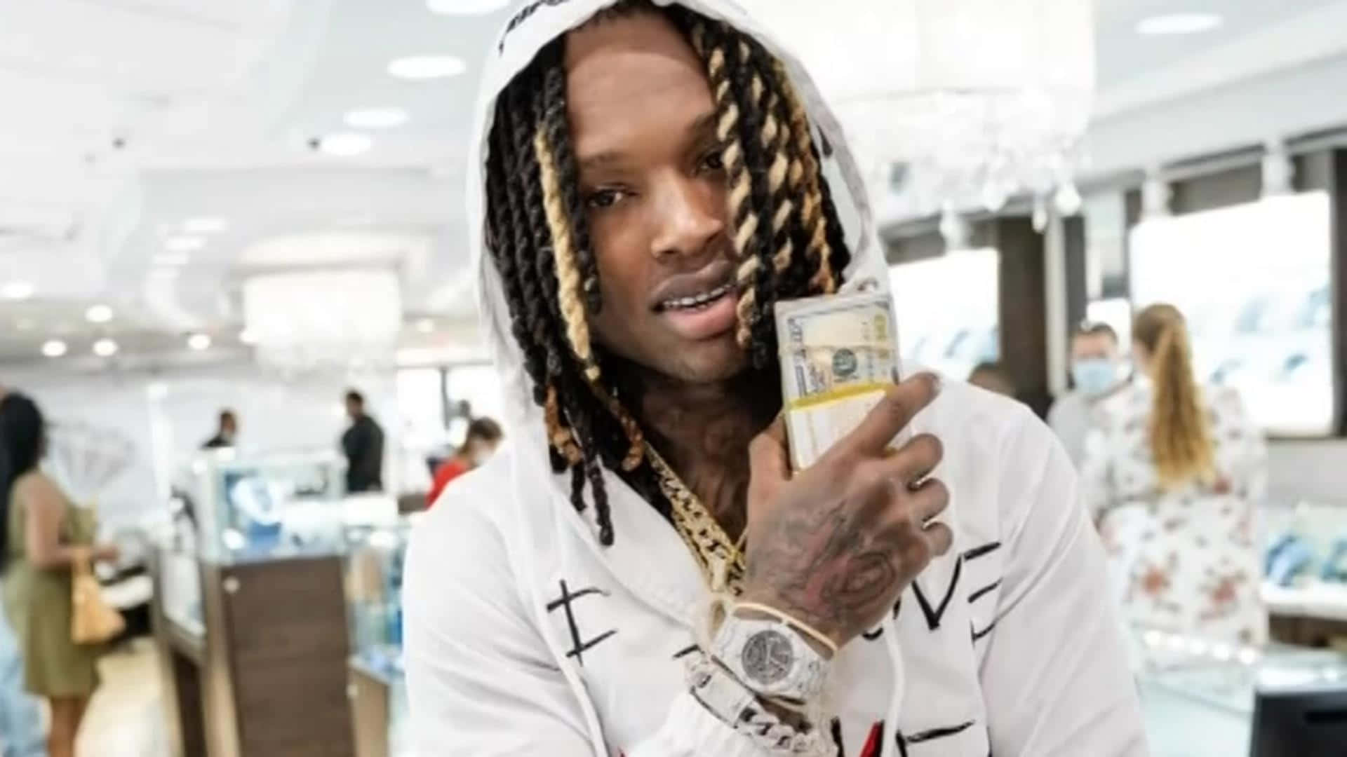King Von 桌布 壁紙 — 一個辮子頭髮的男人穿著白色連帽衫，站在明亮的珠寶店裡，手持一疊現金。