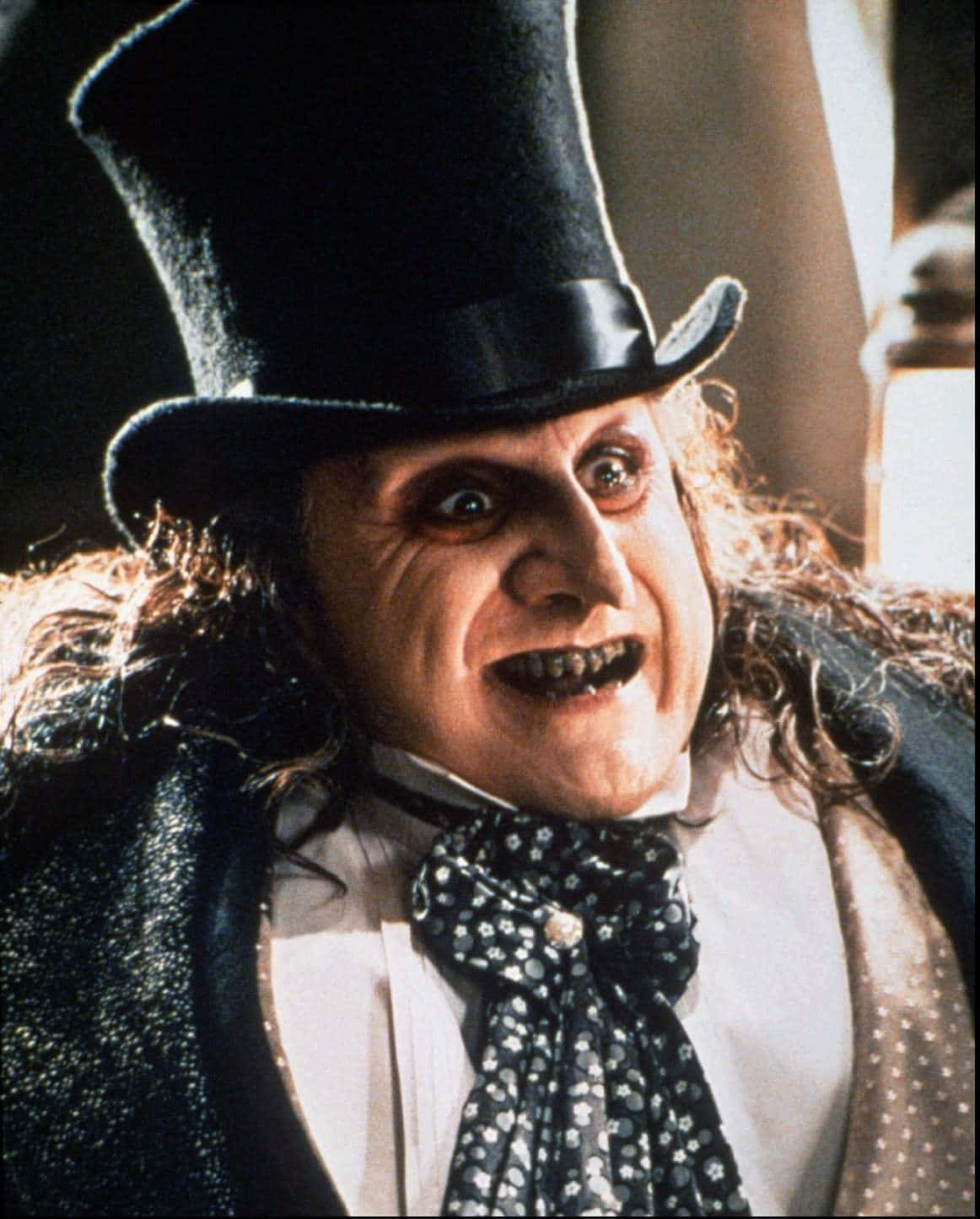 Batman Penguin 桌布 壁紙 — 一個近距離拍攝的邪惡男子，戴著黑色高頂禮帽和圖案領結，露出令人不安的咧嘴笑。