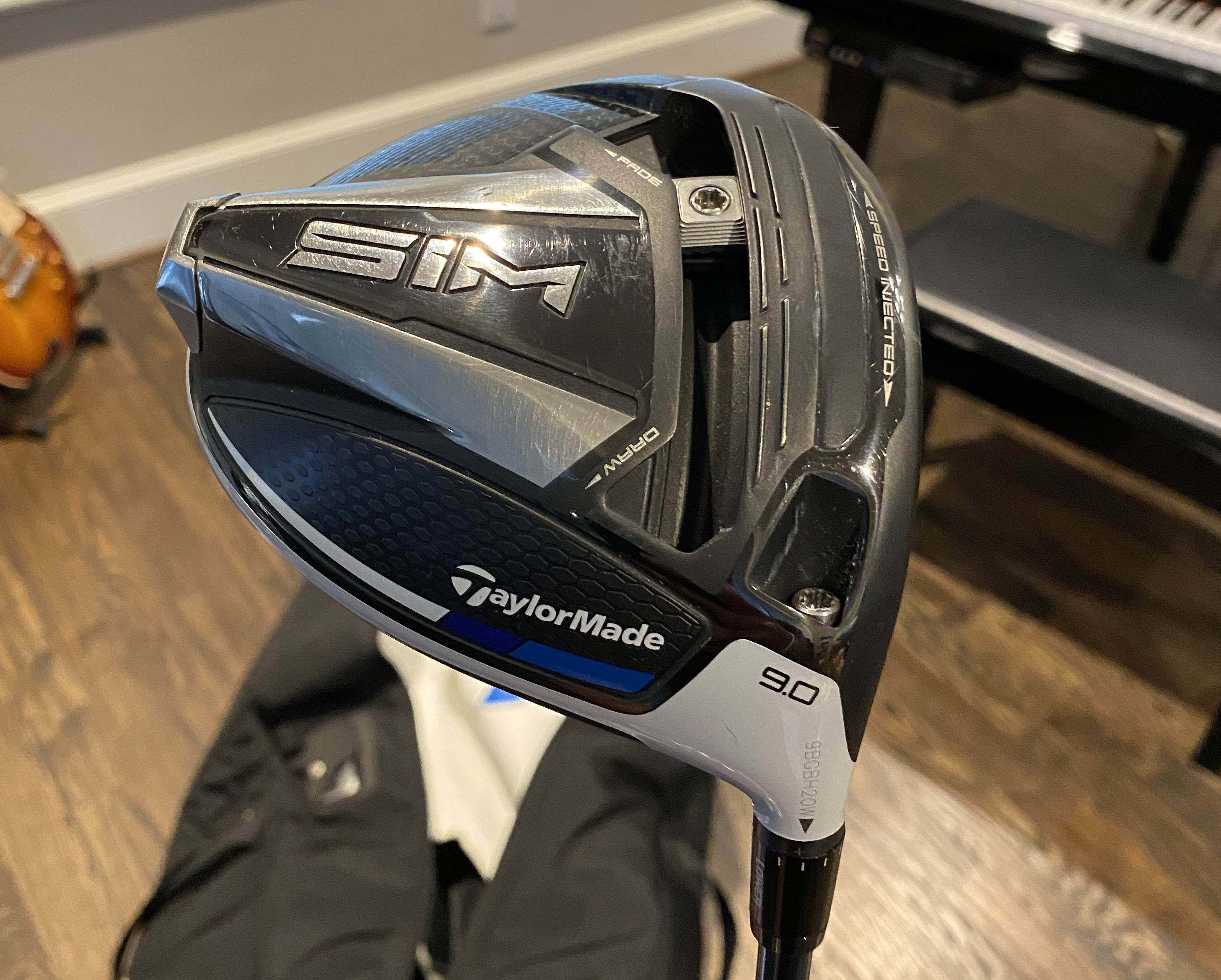 Ryan Palmer 桌布 壁紙 — 一個近距離特寫，展示一支時尚的黑色和銀色TaylorMade SIM高爾夫球桿，桿面傾角為9.0，放置在室內木地板上。