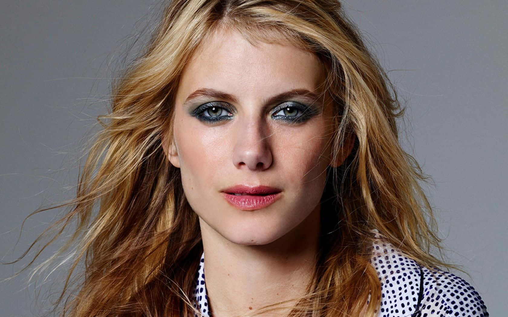 Mélanie Laurent 桌布 壁紙 — 一個近距離肖像，一位女性擁有凌亂的金色頭髮和強烈的煙燻眼妝，背景為中性灰色。