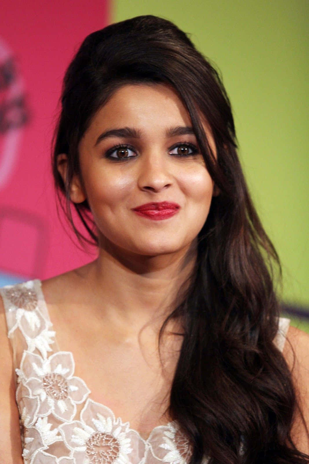 Alia Bhatt 桌布 壁紙 — 一個近距離肖像，一位擁有深色波浪髮和紅色唇膏的女性在充滿活力的粉紅色和綠色背景前溫柔微笑。