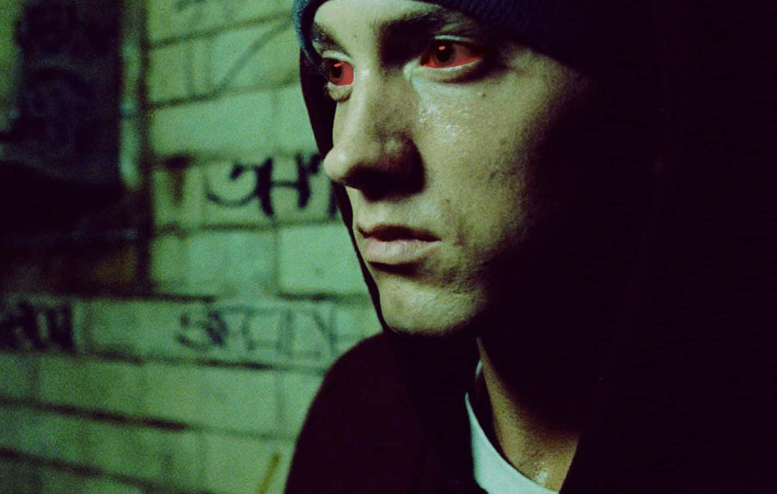 Eminem 桌布 壁紙 — 一個近距離肖像，一個男人在昏暗的綠色光線中，戴著冷帽，眼睛發出紅光，背景是佈滿塗鴉的磚牆。