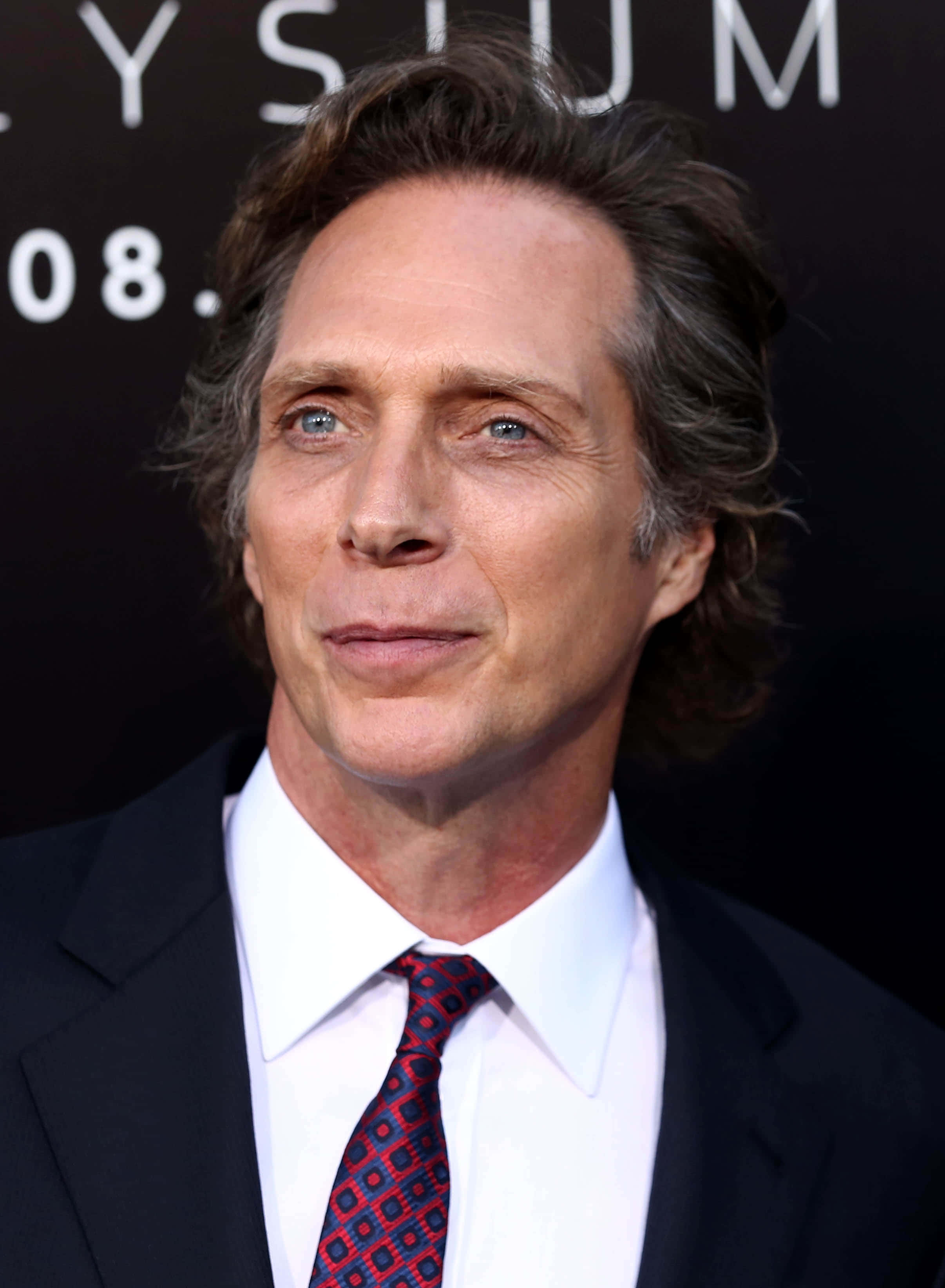 William Fichtner 桌布 壁紙 — 一個近距離肖像，一個男人擁有引人注目的藍色眼睛和波浪棕色頭髮，穿著深色西裝和圖案領帶，背景為黑色並帶有白色文字。