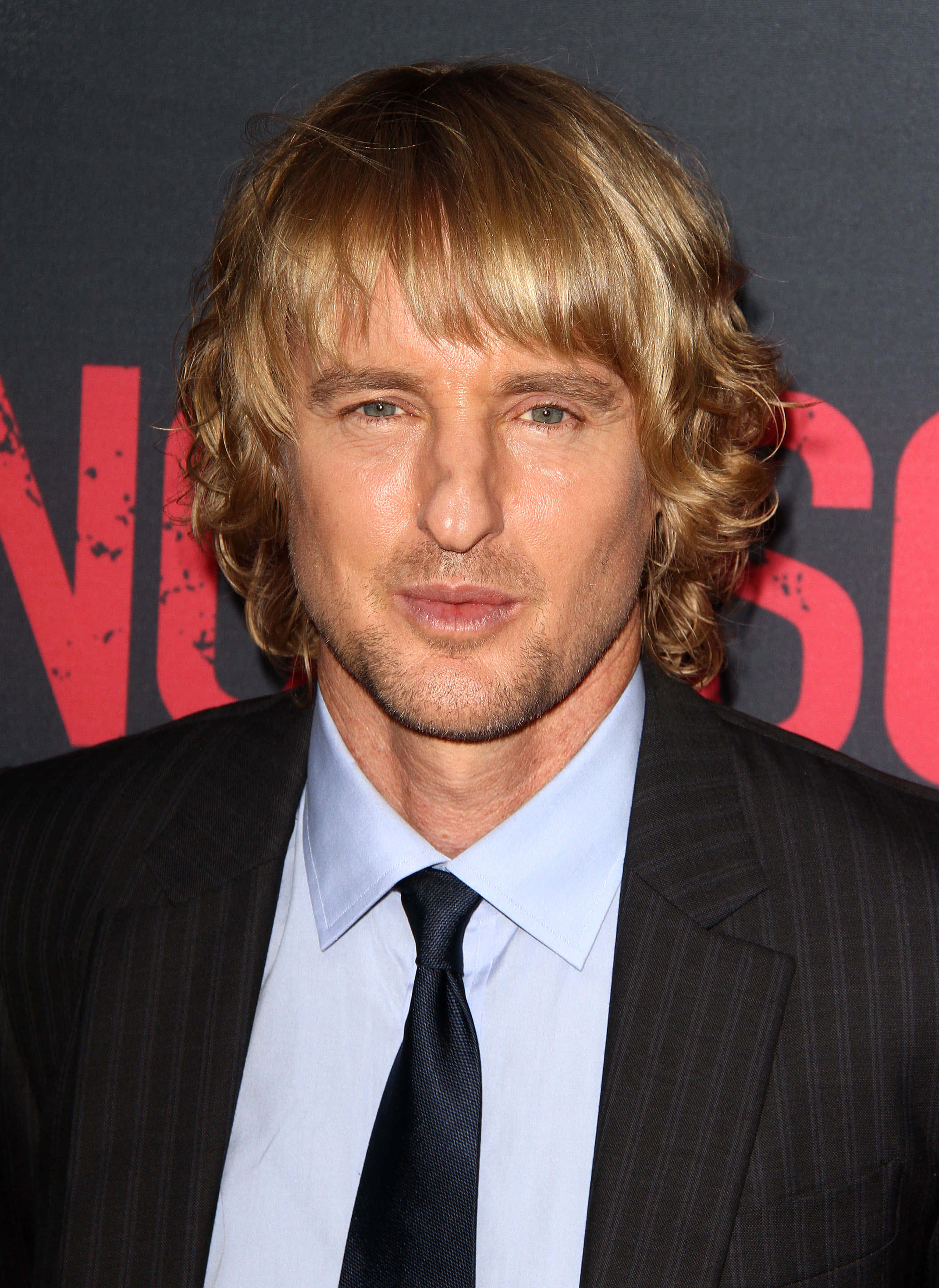 Owen Wilson 桌布 壁紙 — 一個近距離肖像，一名擁有波浪金髮的男子穿著深色細條紋西裝和黑色領帶，背景為深色帶紅色文字。