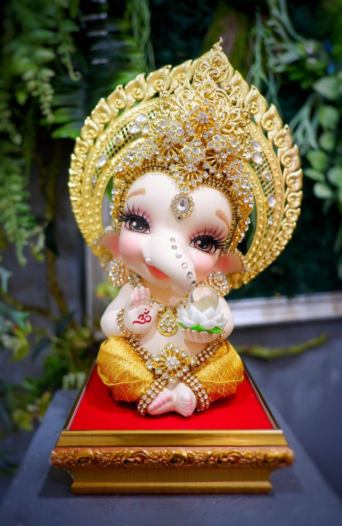 laxmi ganesh saraswati 桌布 壁紙 — 一個迷人風格化的嬰兒象神雕像，飾有黃金珠寶和鑲寶石皇冠，寧靜地坐在紅色軟墊底座上。
