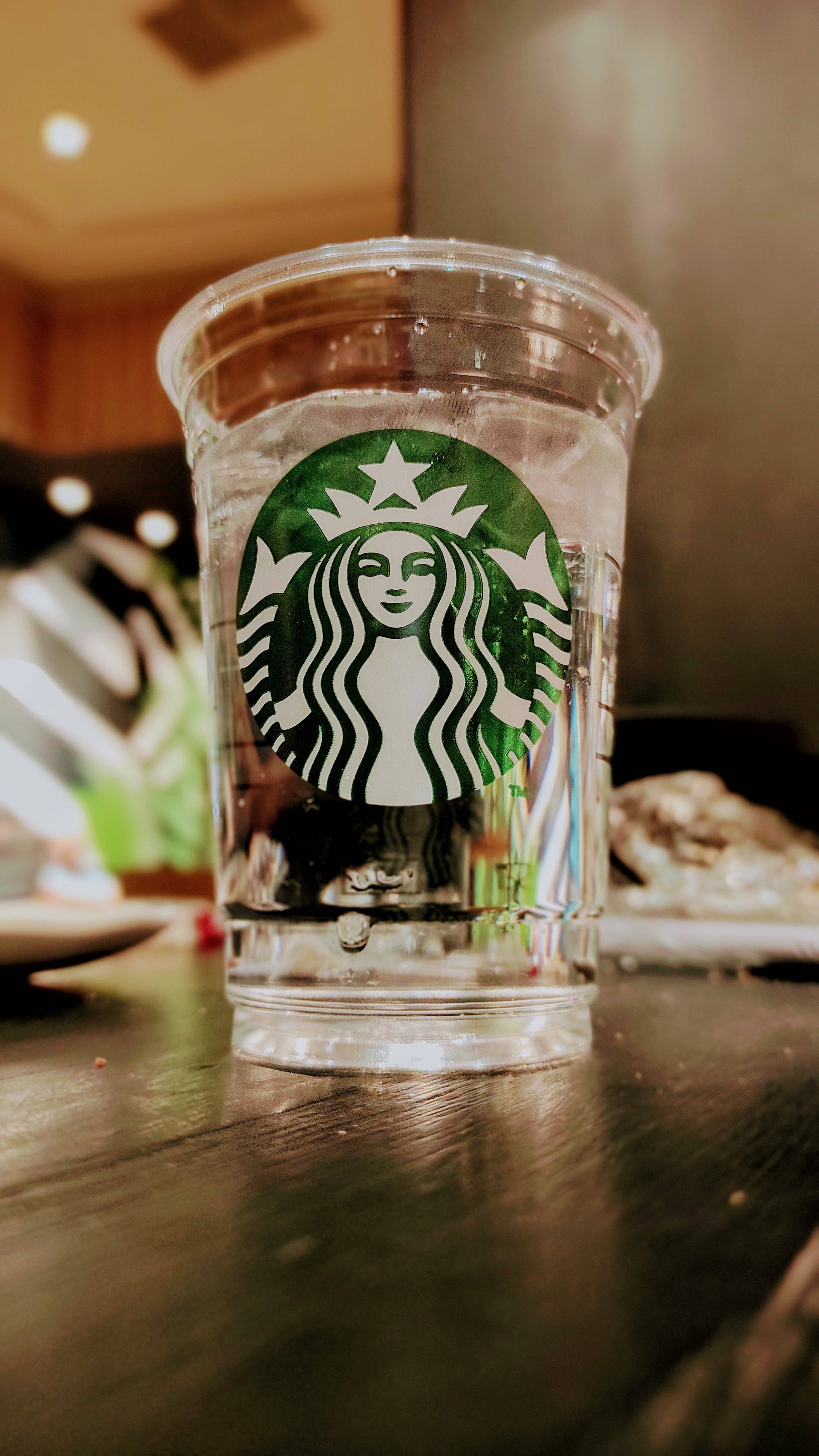 Drink Water 桌布 壁紙 — 一個透明嘅Starbucks杯裝滿水，放喺一間燈光溫暖嘅咖啡店嘅深色木枱上。
