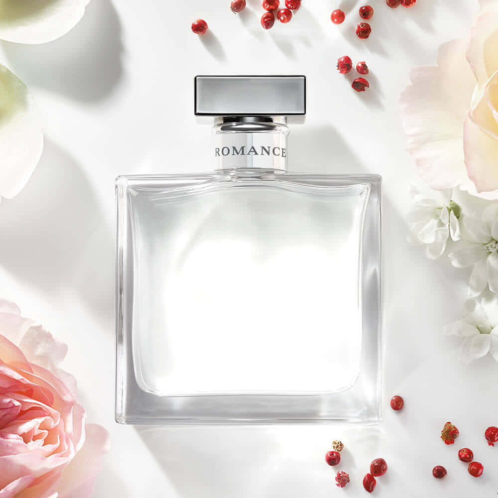 Floral Fragrance 桌布 壁紙 — 一個透明方形香水瓶，標籤上寫著 romance，放在白色表面上，周圍環繞著柔和的粉紅色和白色花朵以及散落的紅色胡椒粒。