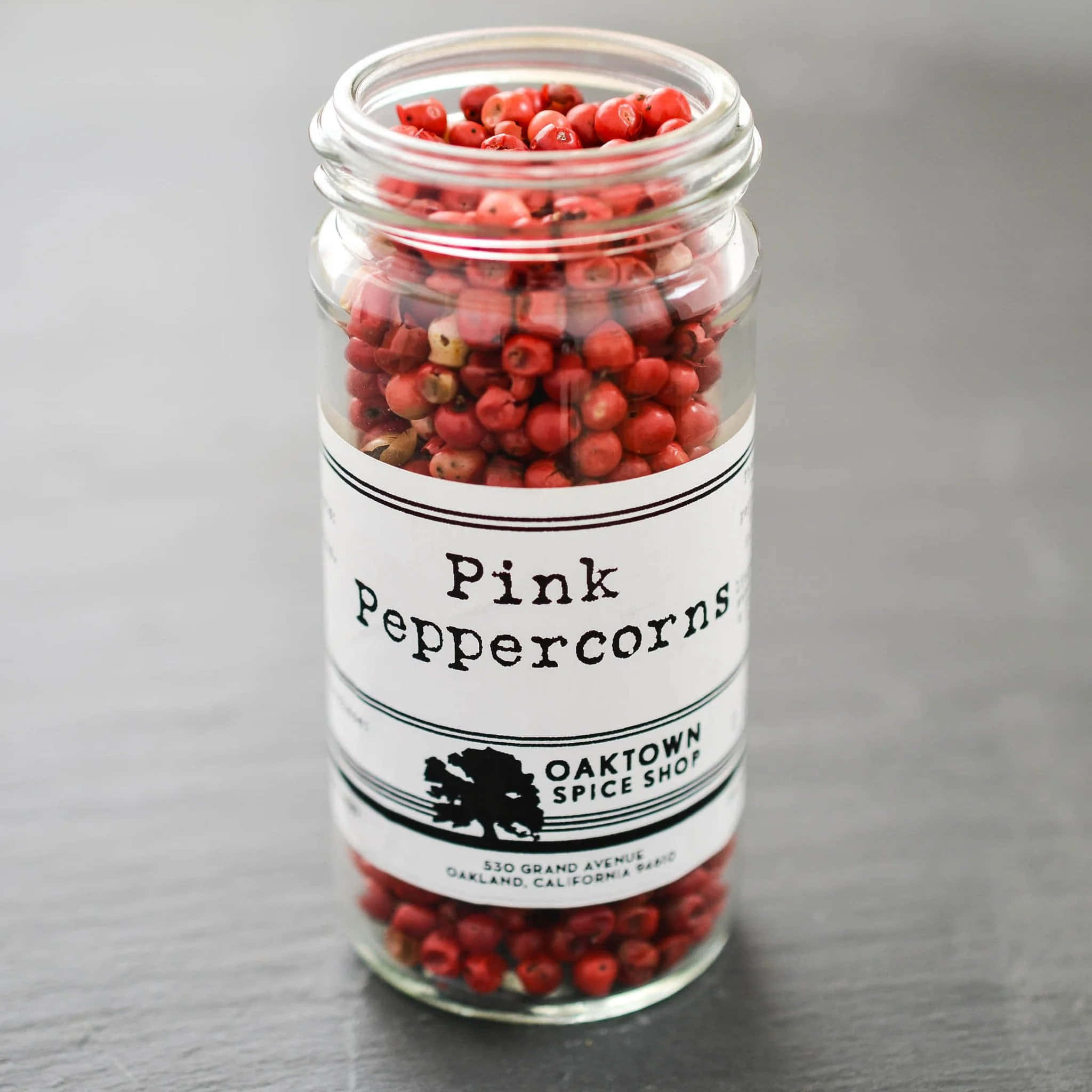 Pink Peppercorn 桌布 壁紙 — 一個透明玻璃瓶裝滿了充滿活力的粉紅胡椒粒，放在灰色石板表面上，帶有極簡標籤，上面寫著來自「Oaktown Spice Shop」的「Pink Peppercorns」。