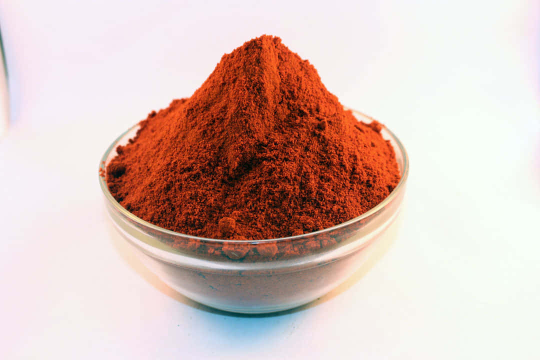 Red Chili Powder 桌布 壁紙 — 一個透明玻璃碗盛載著一堆鮮豔的細磨紅棕色香料，背景是明亮的白色。