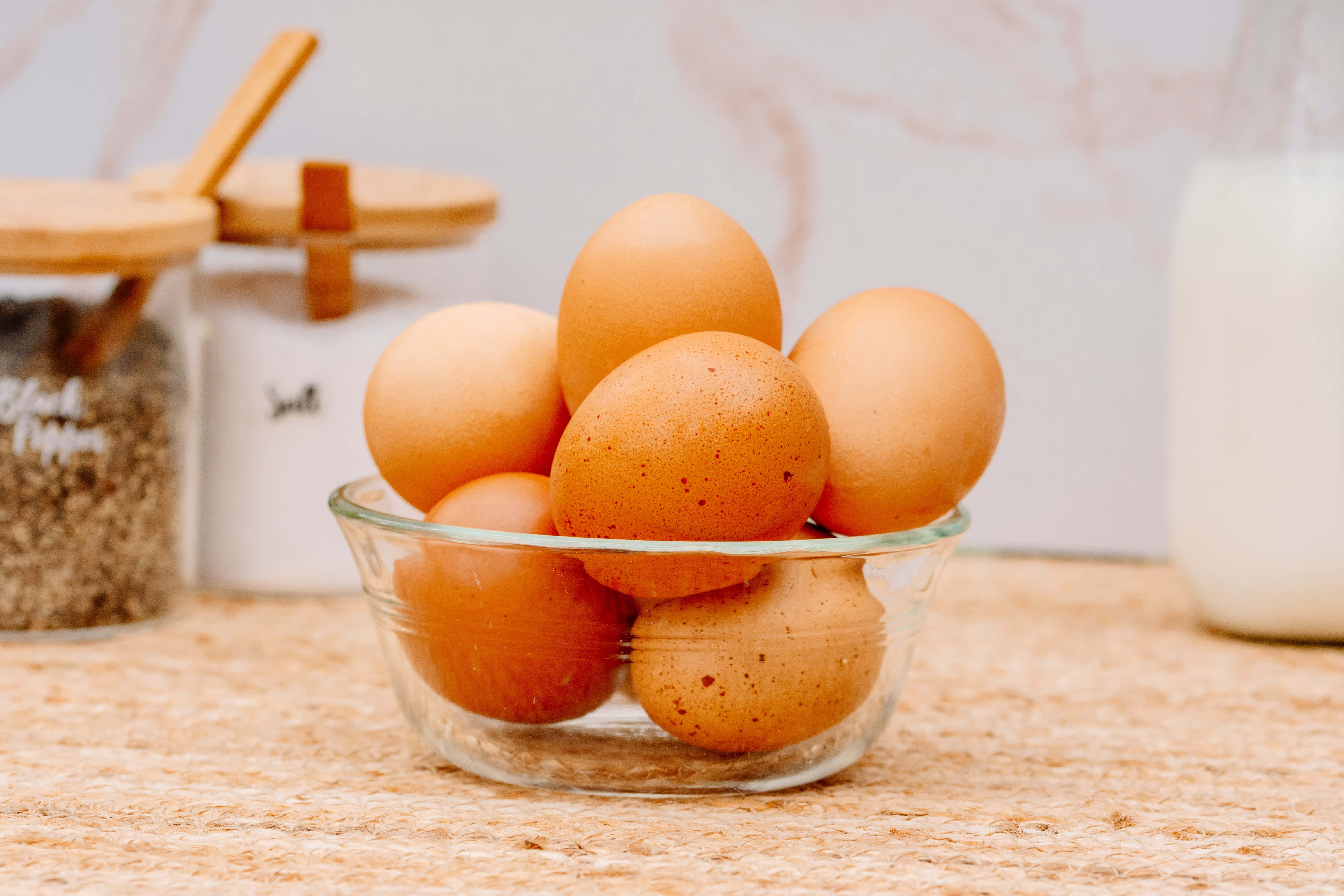 Brown Eggs 桌布 壁紙 — 一個透明玻璃碗裝滿了有斑點的棕色雞蛋，放在編織墊上，旁邊有香料罐和牛奶壺。