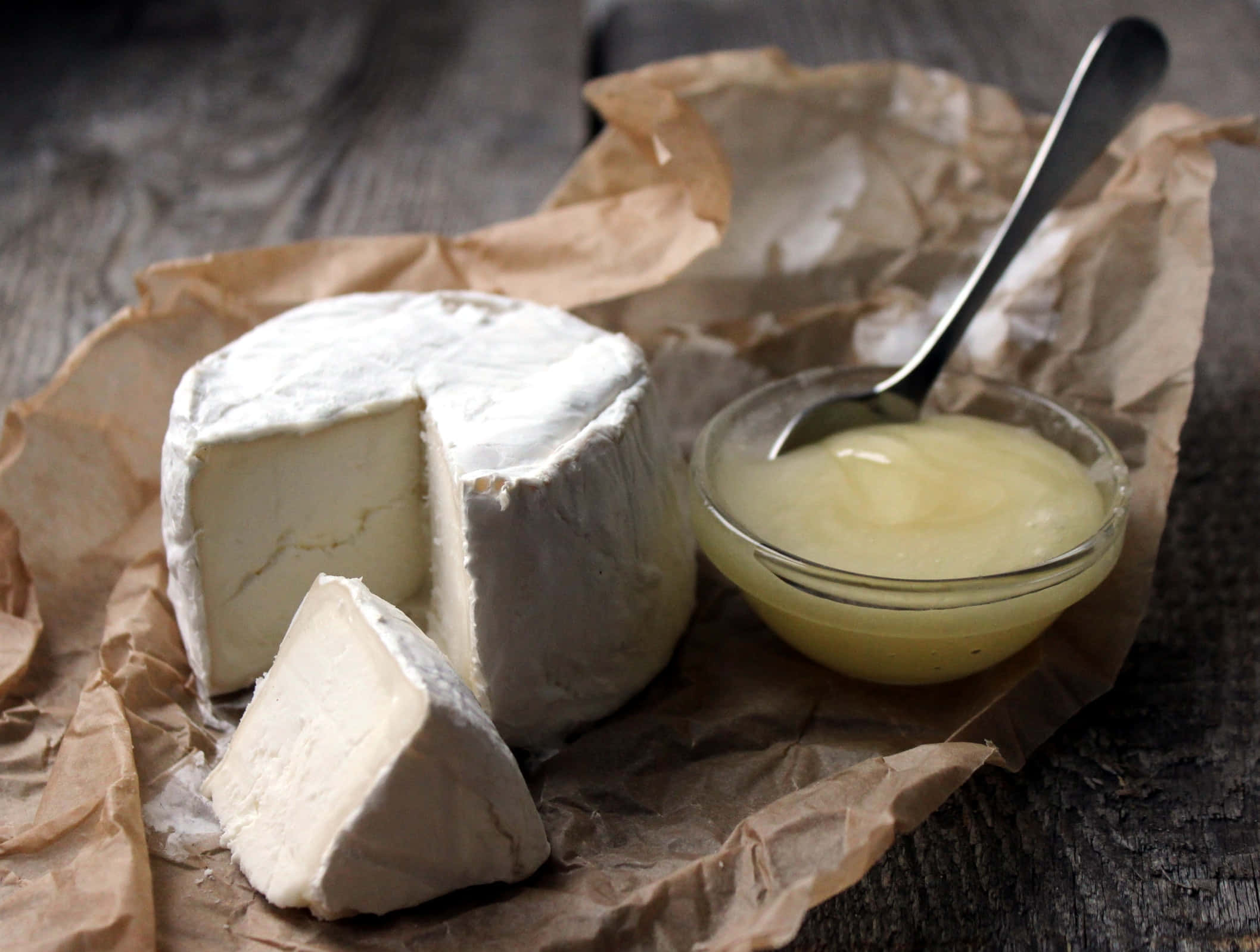 Cheese 桌布 壁紙 — 一個鄉村風格的奶油布里芝士輪，切開一片，蜂蜜放在玻璃碗中，置於皺巴巴的棕色紙上。