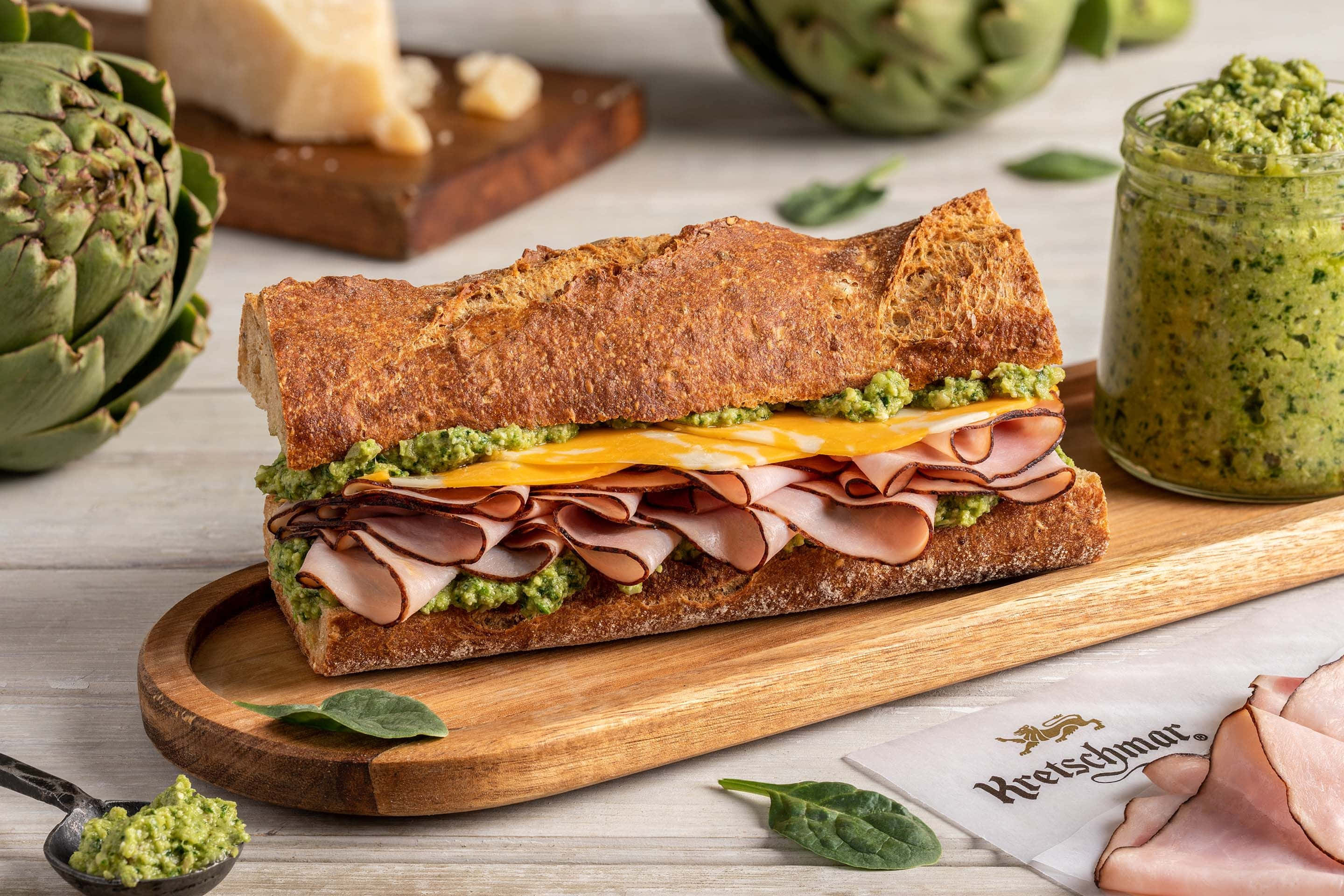 Black Forest Ham 桌布 壁紙 — 一個鄉村風格的帕尼尼，夾著火腿、芝士和香蒜醬，放在木板上，周圍有朝鮮薊和新鮮蔬菜。