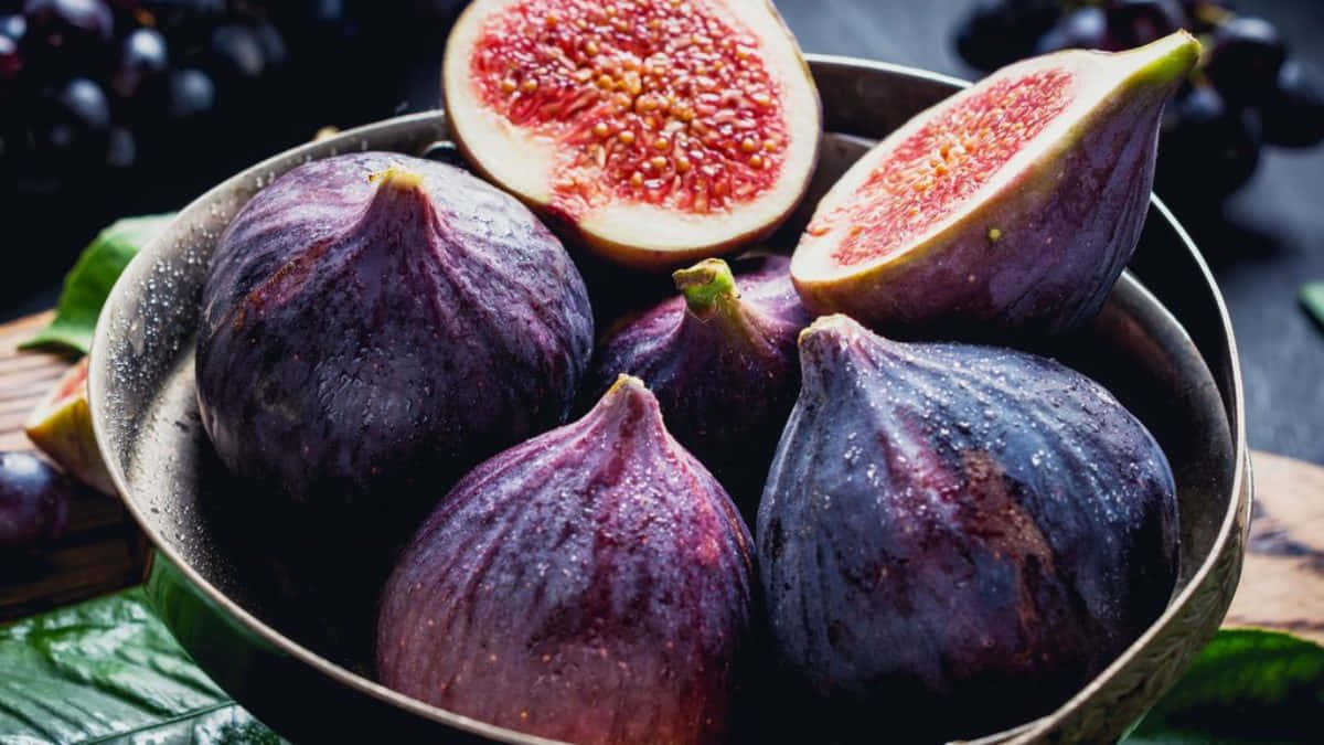 Purple Figs 桌布 壁紙 — 一個鄉村風格的碗盛載著飽滿的紫色無花果，帶有 juicy 的紅色內部和水潤的果皮，襯以深色葡萄和綠葉。