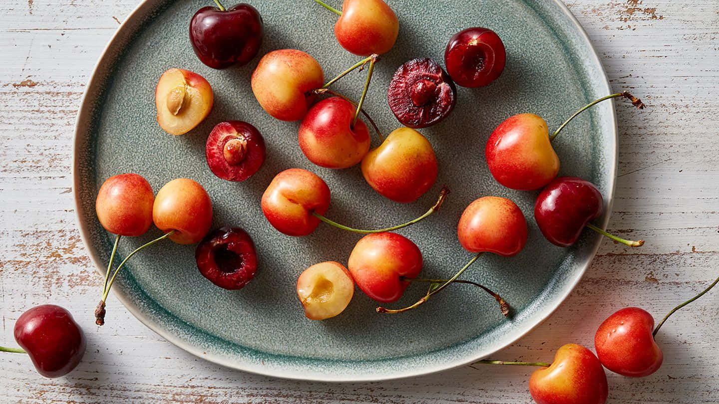 Red Cherries 桌布 壁紙 — 一個鄉村風格的碟子上放著色彩鮮豔的完整和半切櫻桃，置於經風化的白色木質表面上。