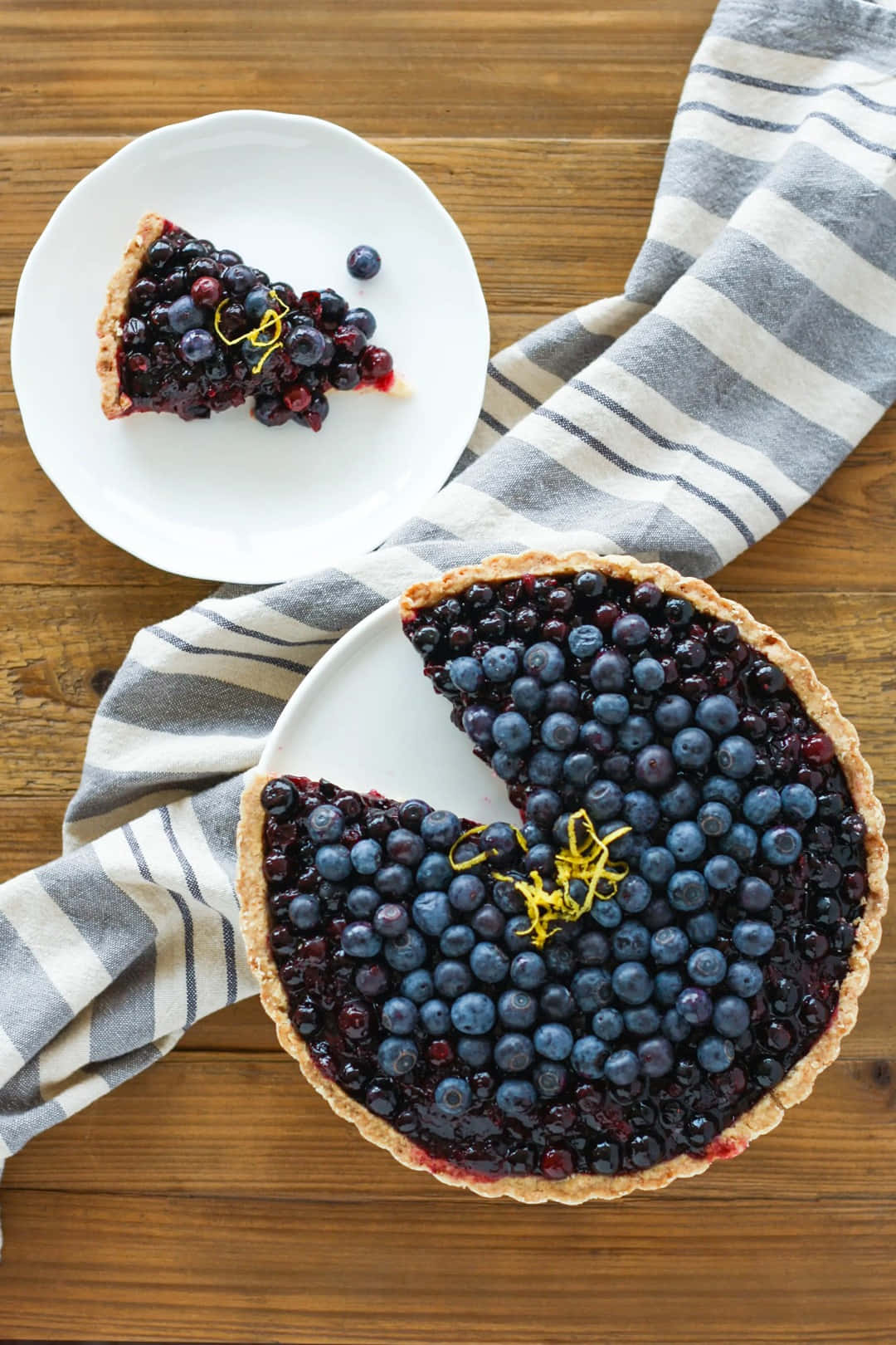 Blueberries Tart 桌布 壁紙 — 一個鄉村風格的藍莓撻，切片放在白色碟子上，旁邊有條紋布，置於木桌上。