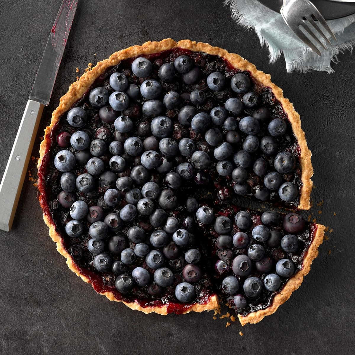 Blueberries Tart 桌布 壁紙 — 一個鄉村風格的藍莓撻，金黃色的外皮，放在深色石板表面上，附近有刀和叉。