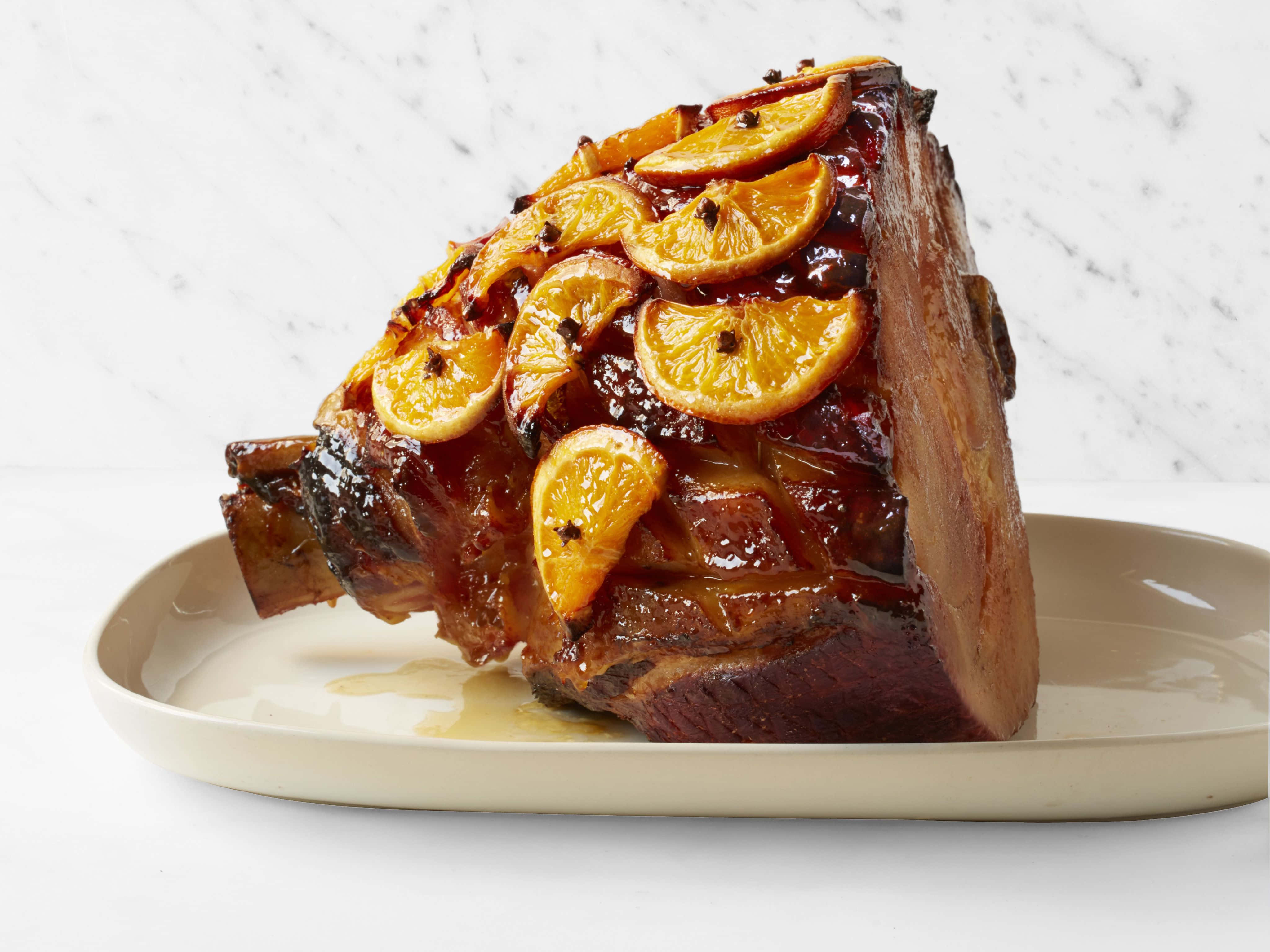 Black Forest Ham 桌布 壁紙 — 一個釉面火腿，上面放有蜜餞橙片和整粒丁香，放在米色碟子上，背景是白色大理石。