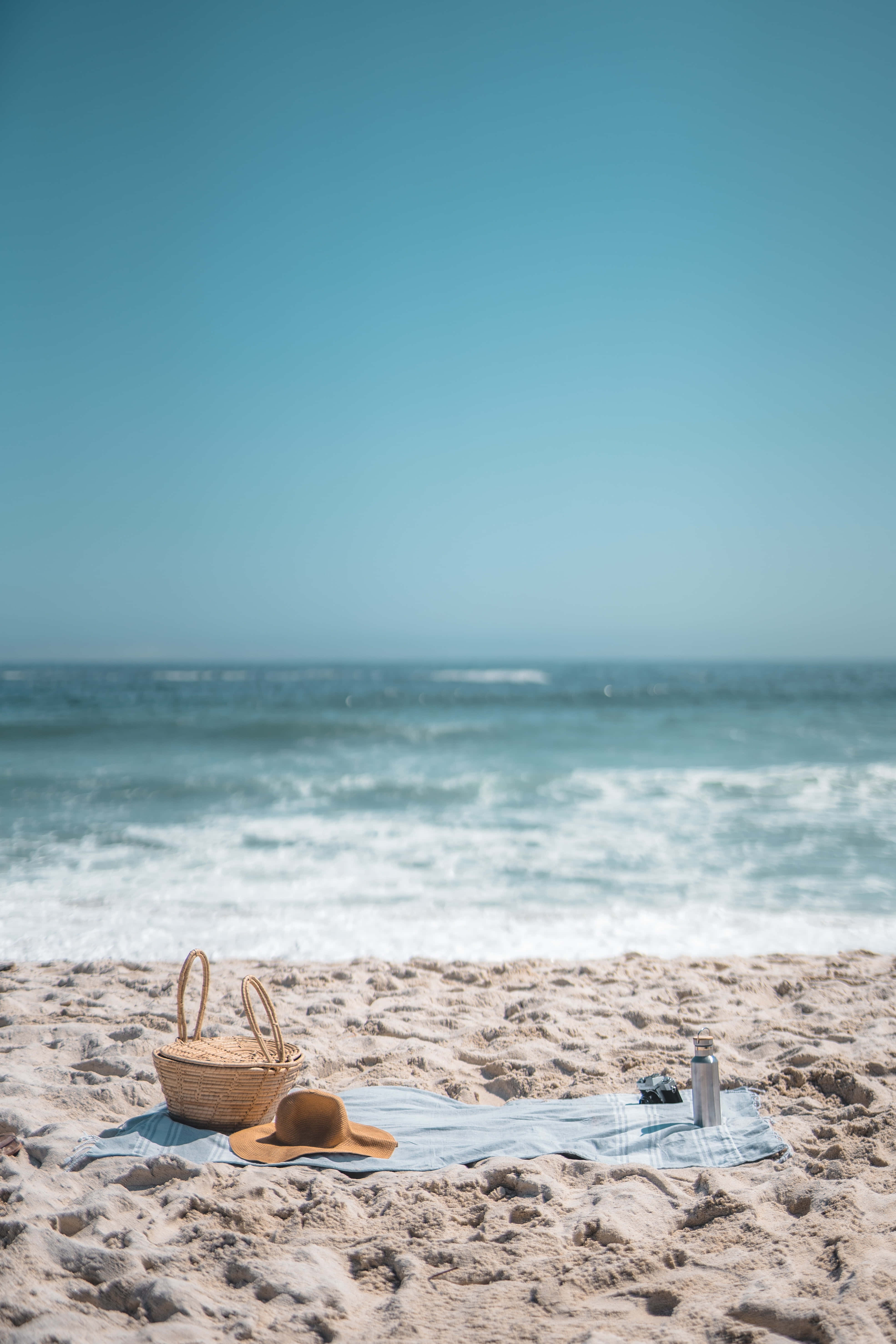 Beach Picnic 桌布 壁紙 — 一個野餐佈置，藤籃和草帽放在藍色毛巾上，毛巾置於陽光普照的沙灘上，旁邊是海洋，上方是清澈的藍天。
