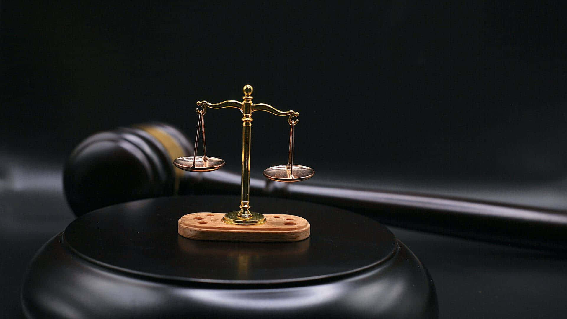 Justice 桌布 壁紙 — 一個金色微型正義天秤居中放置在帶有木質底座的黑色槌頭上，背景為深色。