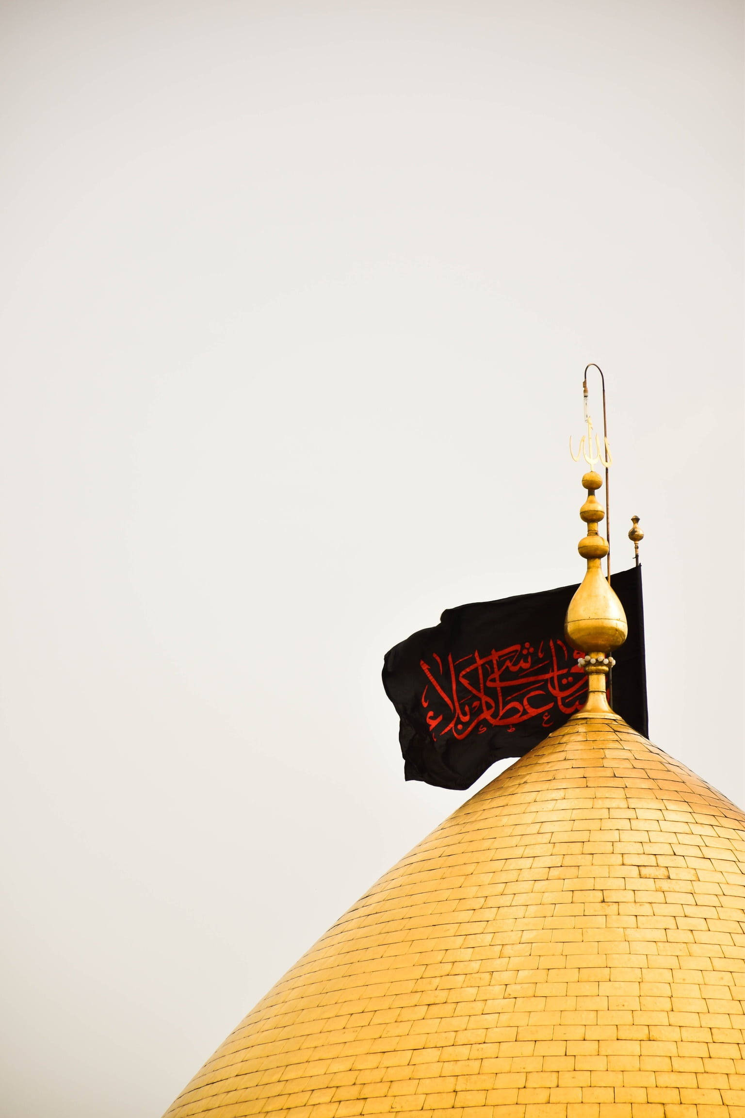 Karbala 桌布 壁紙 — 一個金色瓷磚圓頂，頂部有一面黑色旗幟，上面有紅色阿拉伯書法，襯著蒼白的天空。