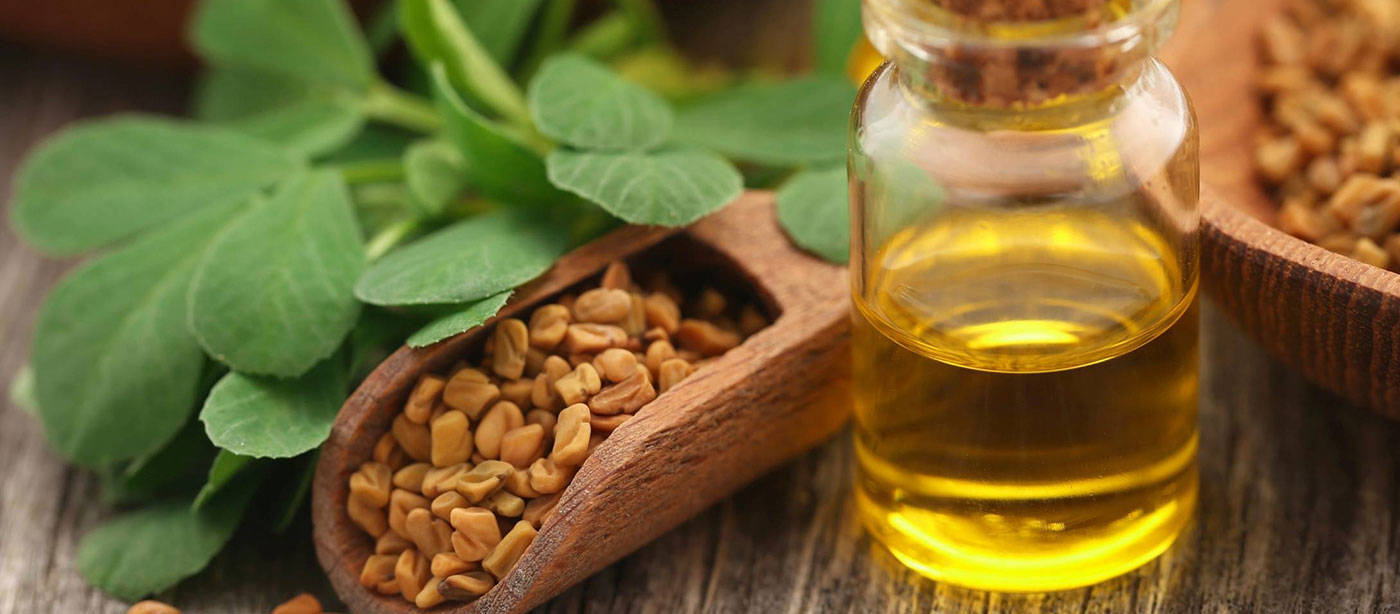 Fenugreek Oil Bottle 壁紙 — 一個金色葫蘆巴油的玻璃瓶旁邊放著裝滿種子和新鮮綠葉的木勺，置於質樸的木質表面上。