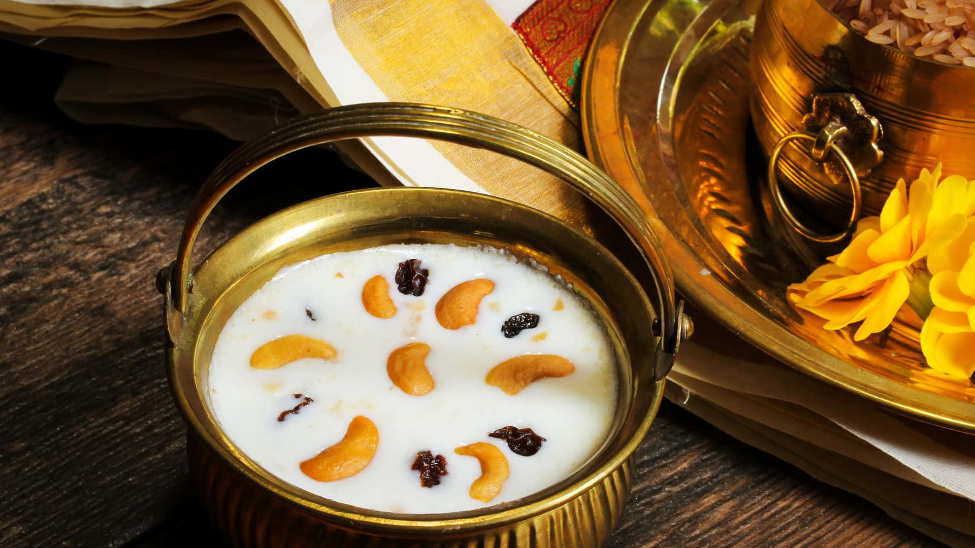 Vishu 桌布 壁紙 — 一個金色黃銅碗盛載著奶油白色的卡爾，上面放有腰果和提子乾，旁邊有一朵黃花和傳統紡織品。