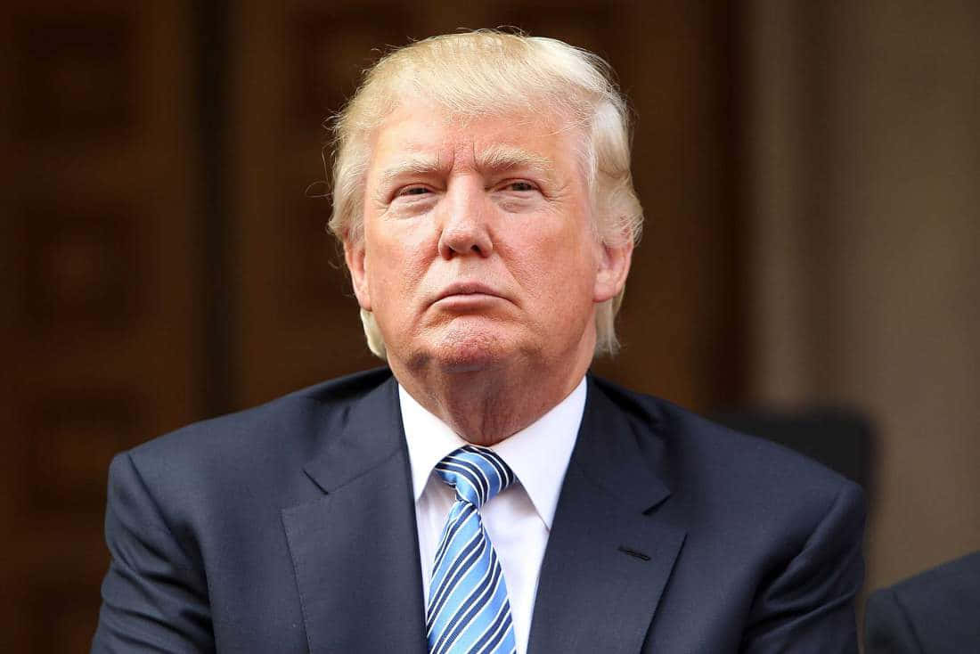 trump 桌布 壁紙 — 一個金髮男士的特寫肖像，穿著深色西裝和藍色條紋領帶，背景是模糊的木質背景。